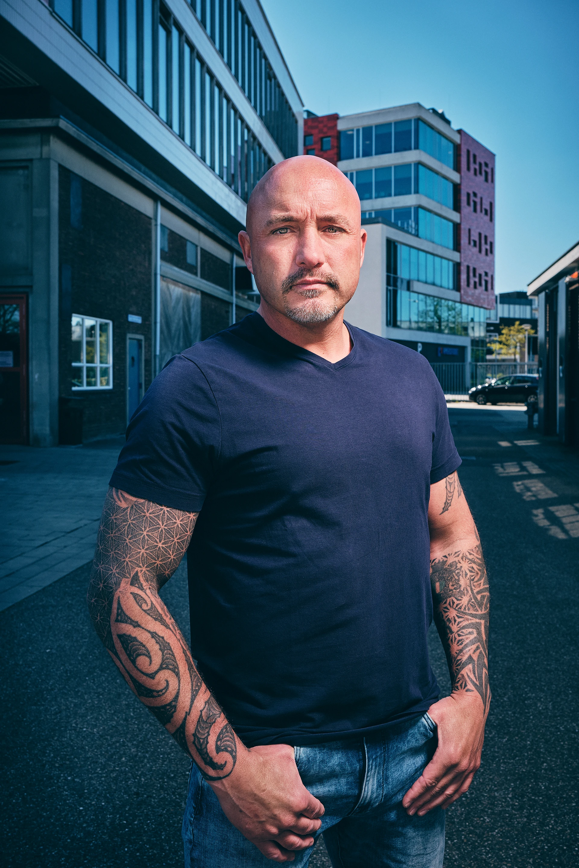 Bald man with tattoos on both arms wearing a navy shirt and jeans standing outdoors in an urban area.