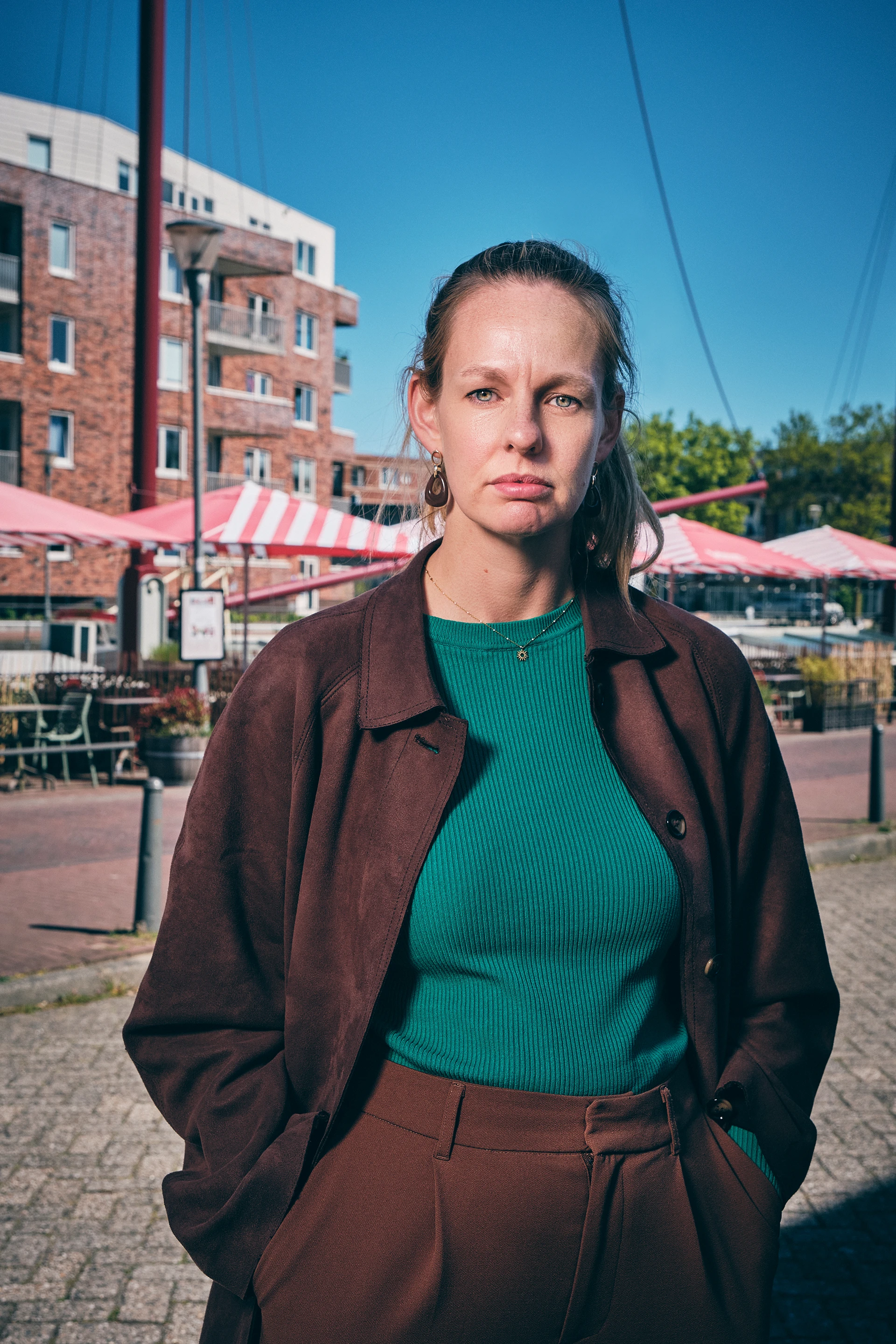 Woman in teal sweater and brown coat standing outdoors near brick buildings and striped tents.