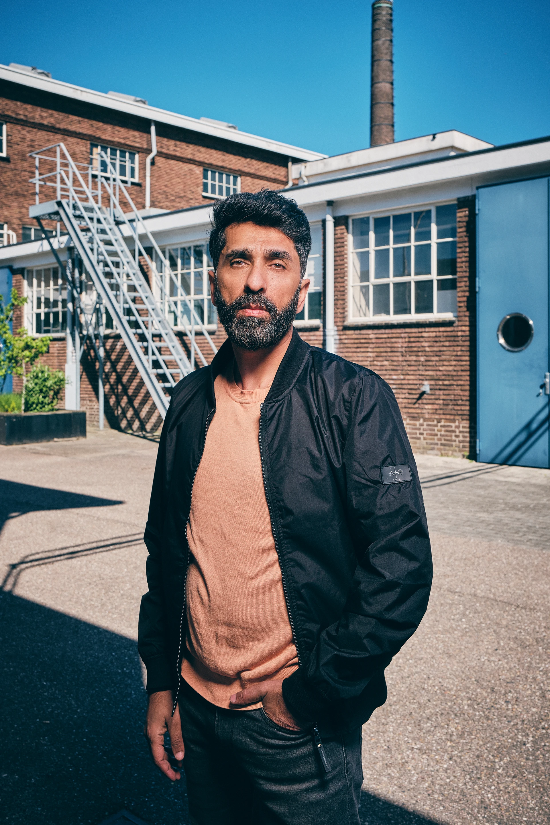 Man with dark hair and beard wearing black jacket and tan sweater outside brick building near stairs.