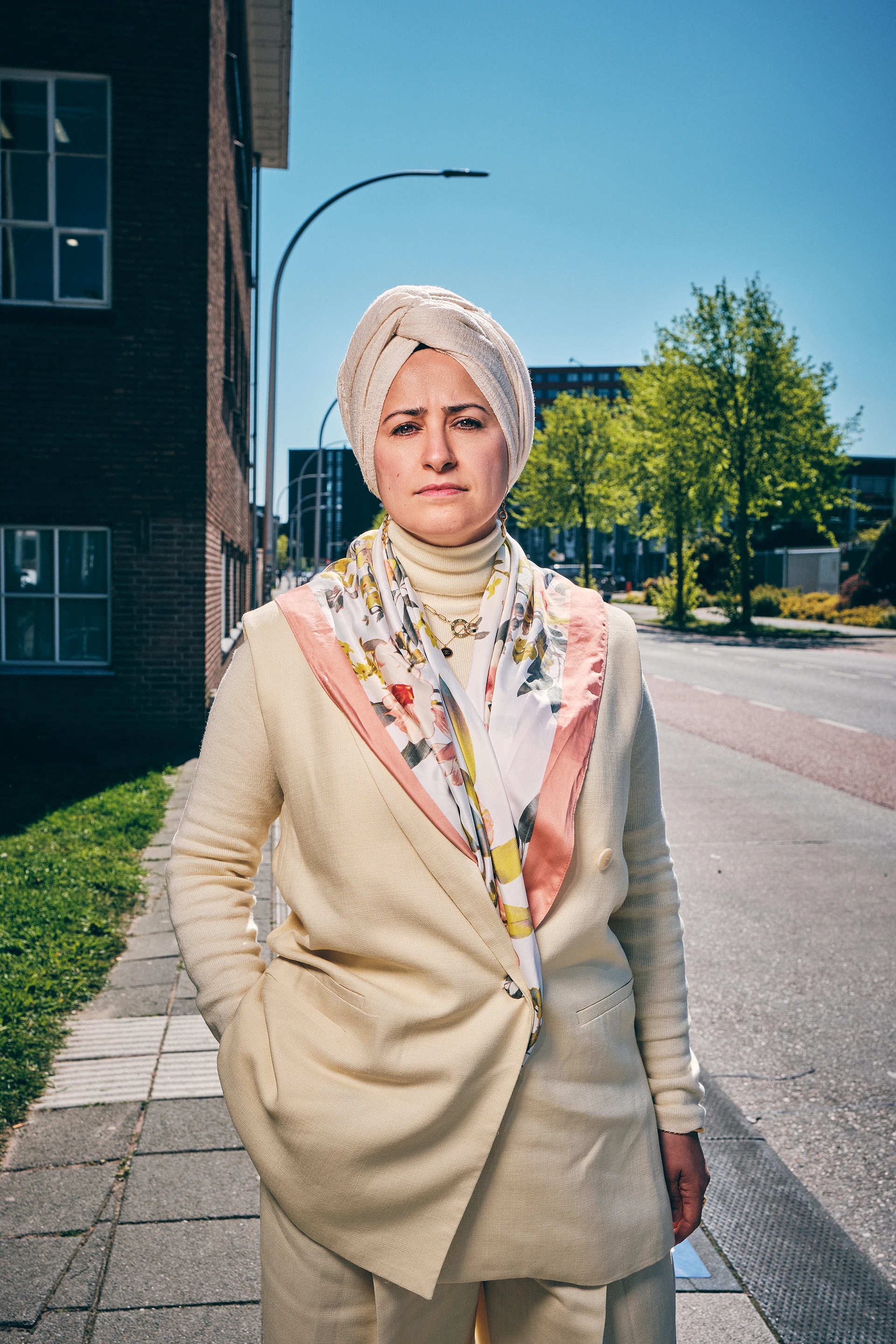 Woman wearing a beige turban and suit with floral scarf standing on a city sidewalk on a sunny day.