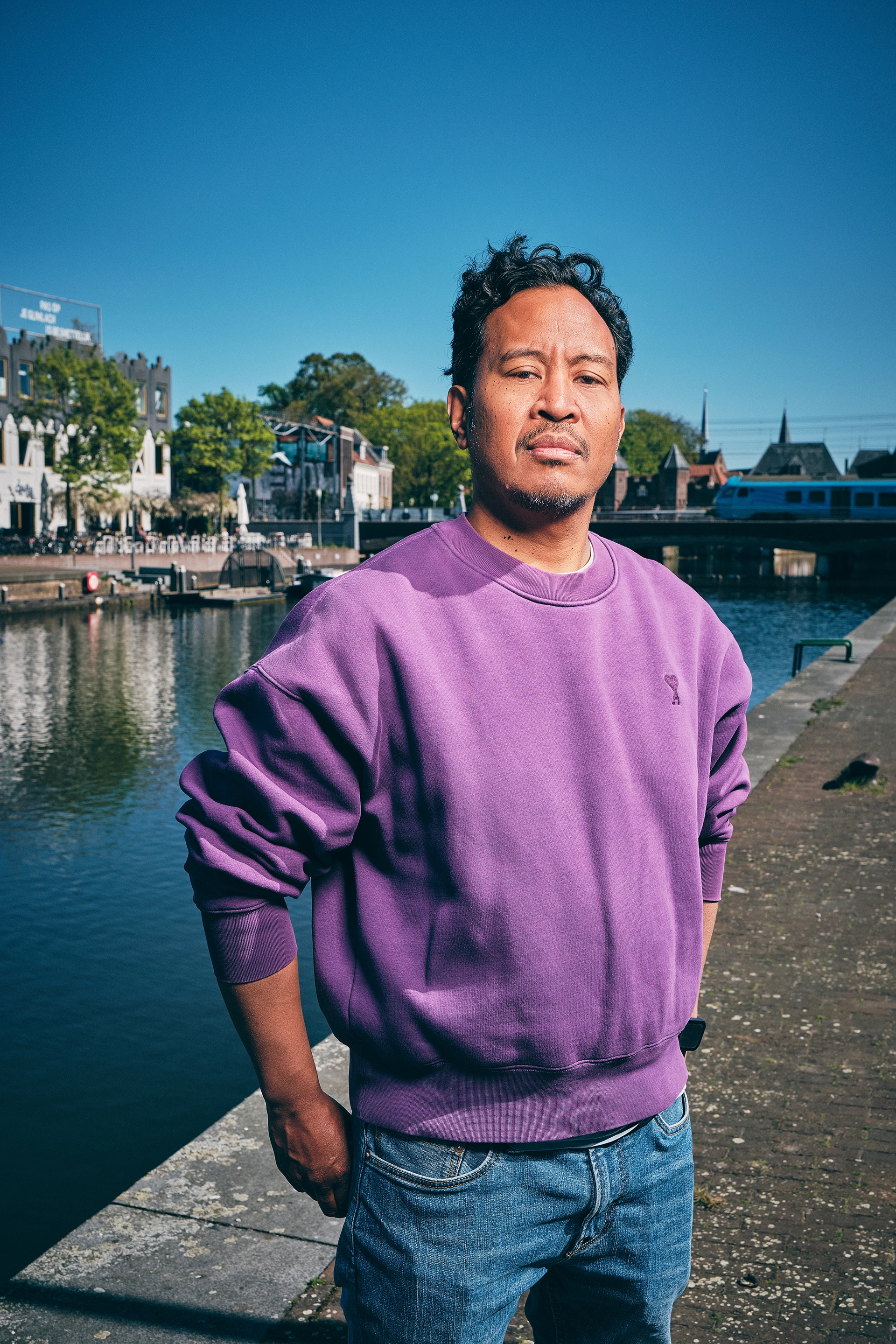 Man in a purple sweatshirt standing confidently by a canal on a sunny day with blue sky.
