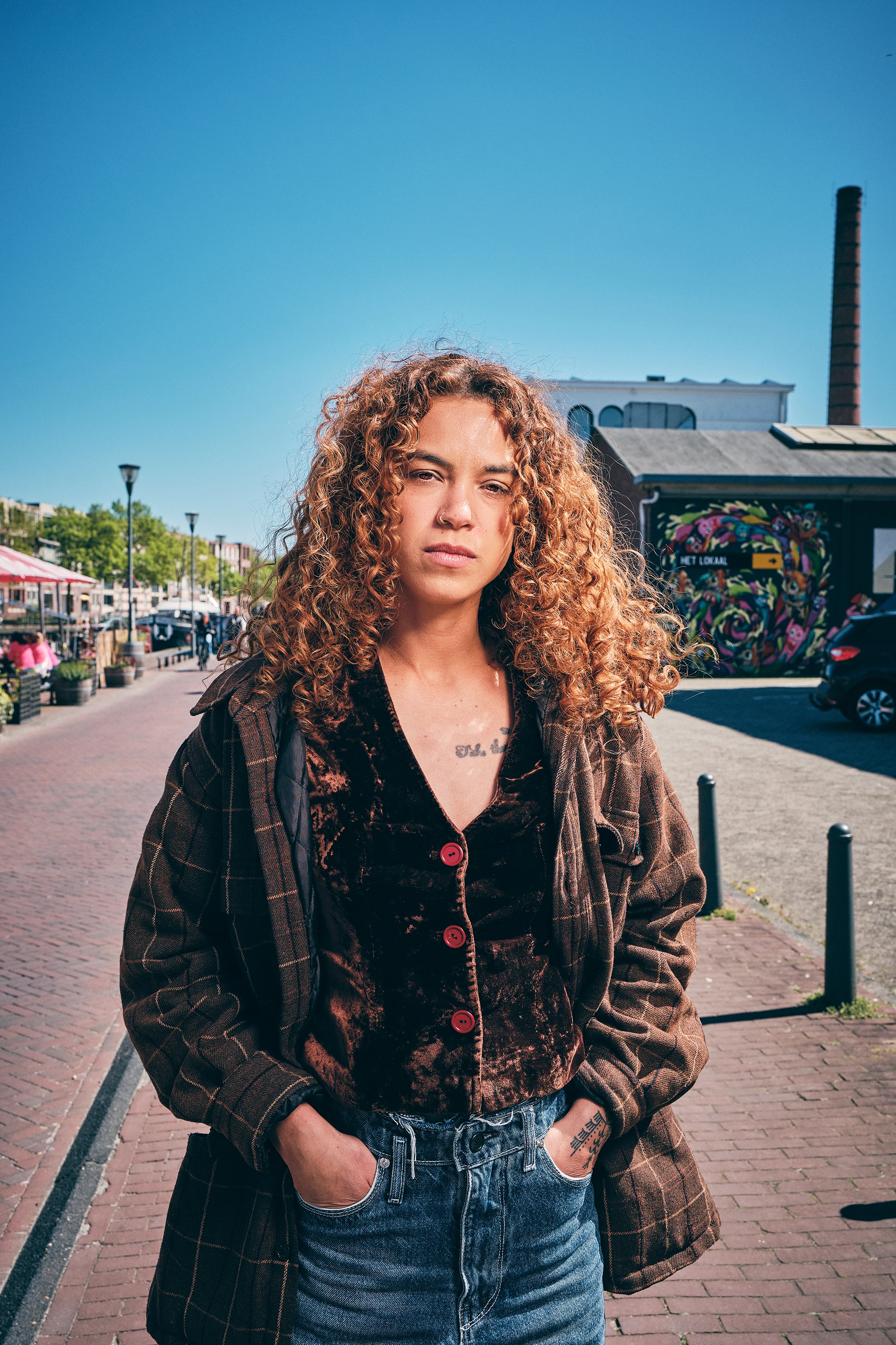 Woman with curly hair, wearing a brown jacket and velvet top, standing on a sunny urban street.