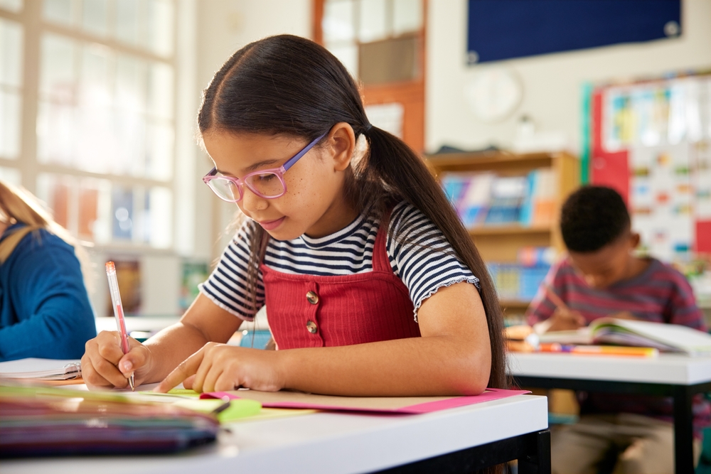 Concentrated pupil at the desk is writing in book with classmates in classroom.