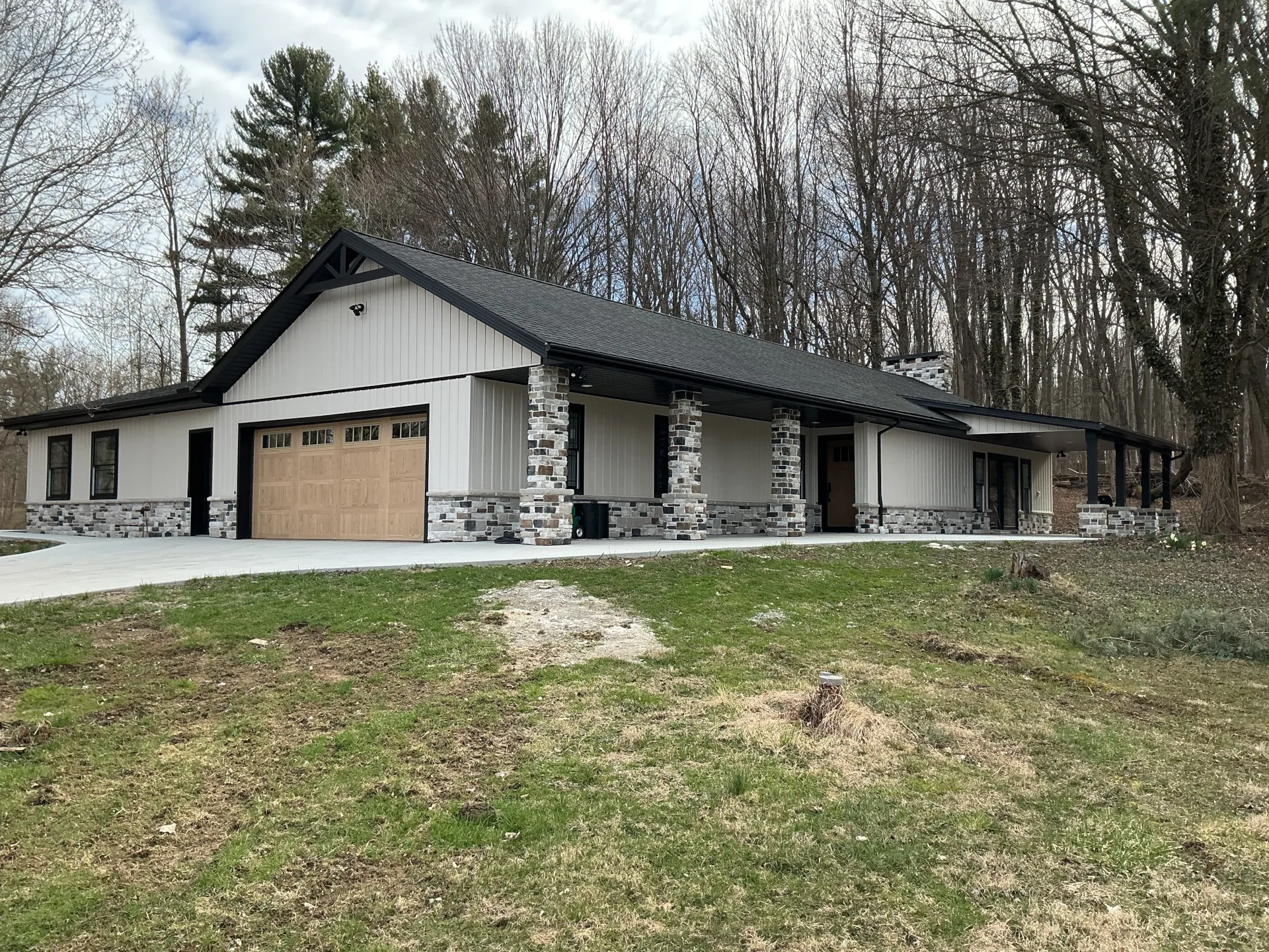 The angled view of a house with a wooden garage door, stone veneer base, grey vinyl siding, and a black roof.