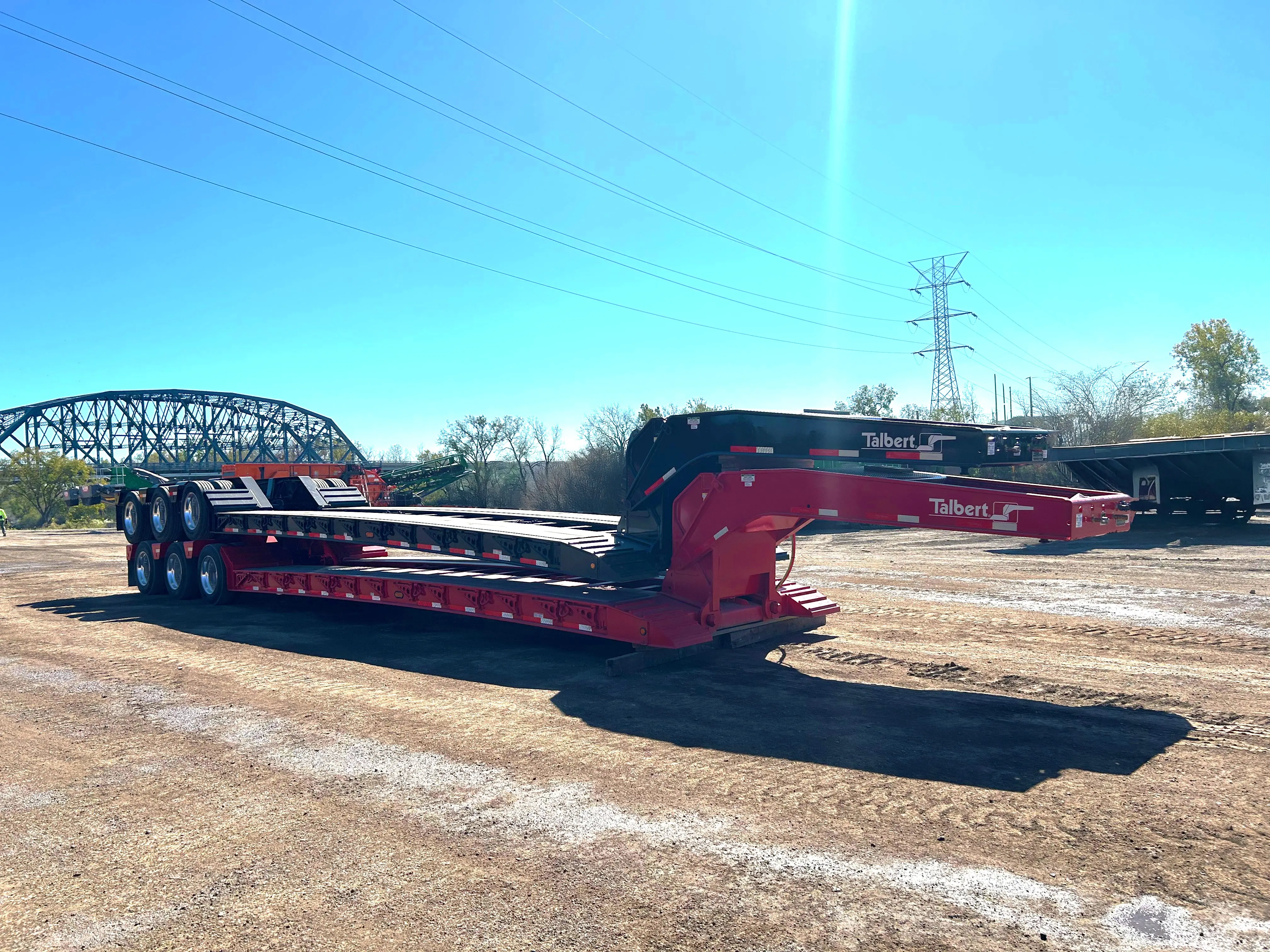 A large red Talbert lowboy equipment transport trailer with multiple axles, designed for hauling heavy machinery, parked on a dirt lot beneath a sunny sky.