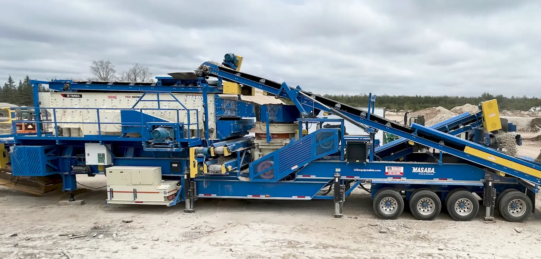A long, low-profile blue track-mounted mobile aggregate crusher or screener (McCloskey or similar) with multiple conveyors, operating at a quarry or crushing site.