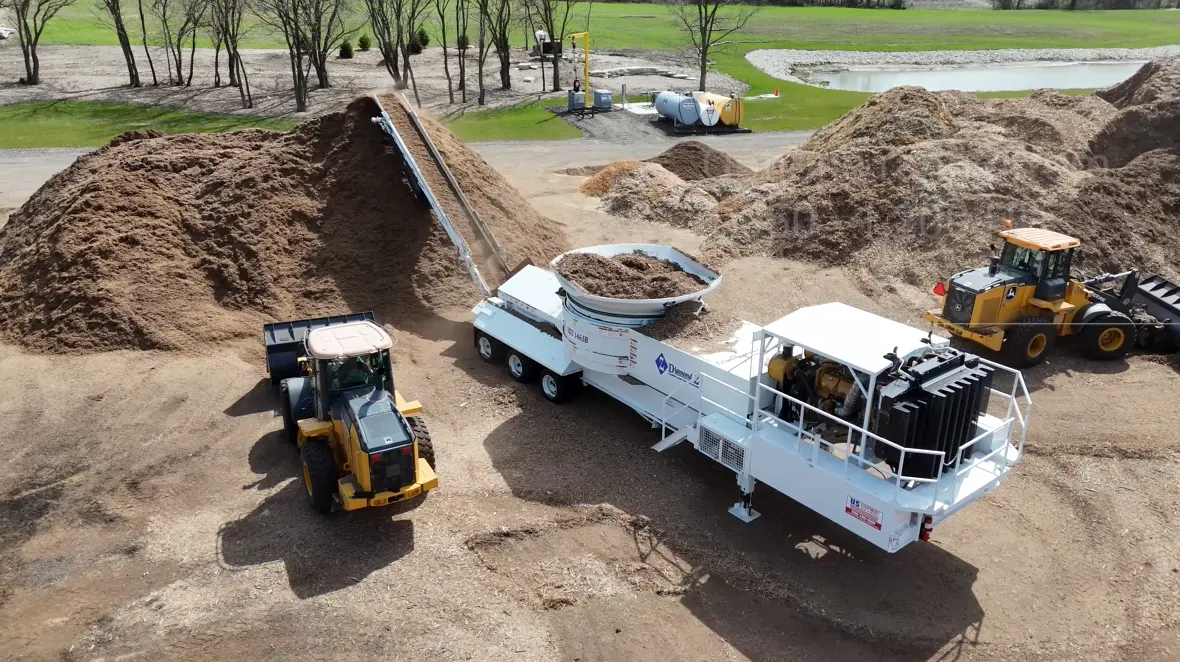 A white and black stationary or portable trommel screen and trommel feed hopper processing a large pile of wood mulch or aggregate material, with yellow heavy loaders nearby.