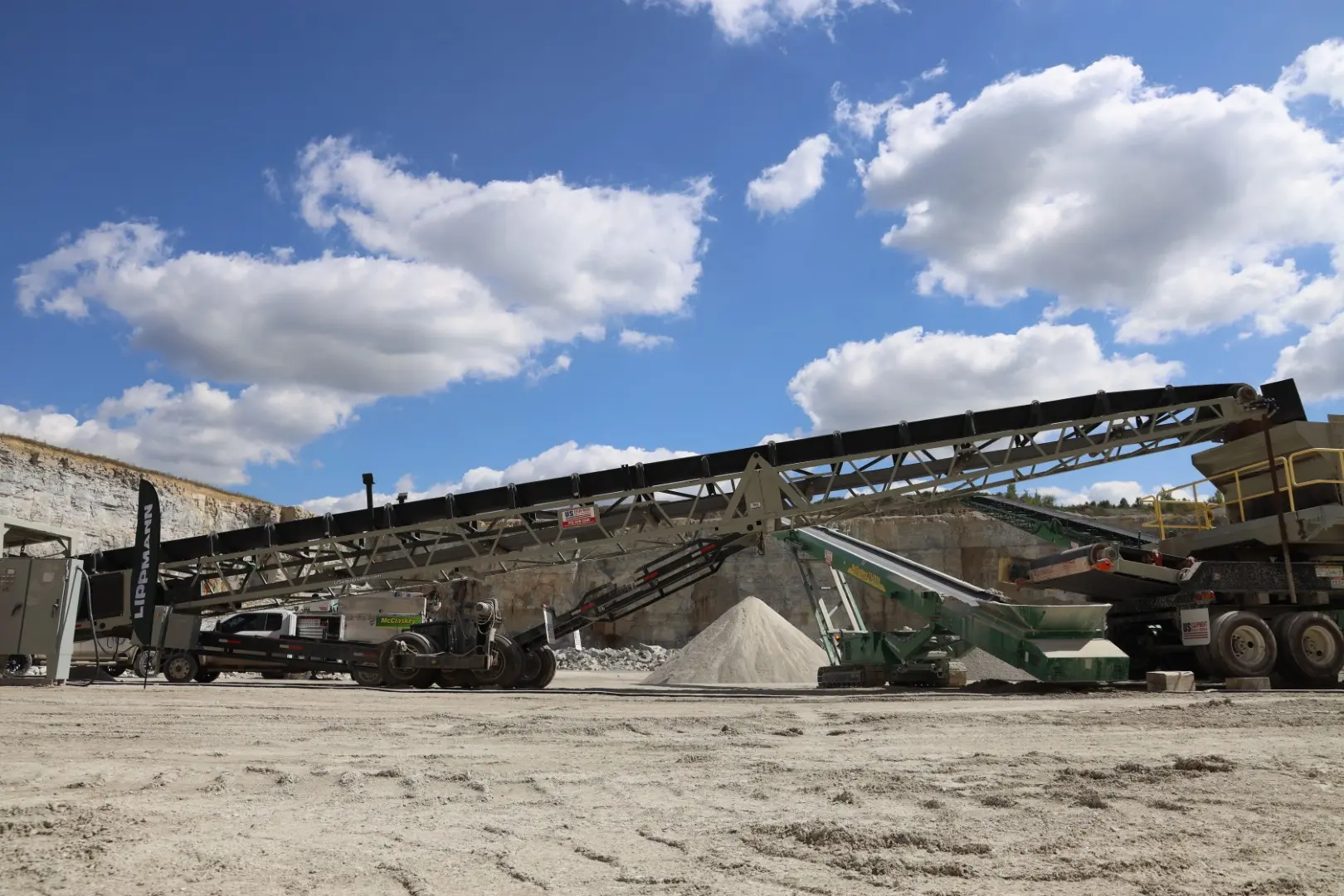 A portable, track-mounted crushing or screening plant operating near a quarry wall, featuring a long conveyor belt extending up and over a pile of sand or fine aggregate.