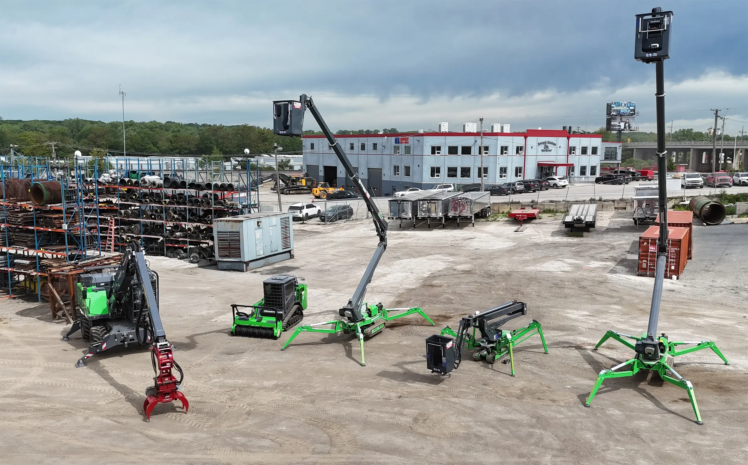 An outdoor equipment lot showing specialized compact machinery with green legs (spider lifts or micro excavators) and various attachments in the foreground of a large warehouse building.