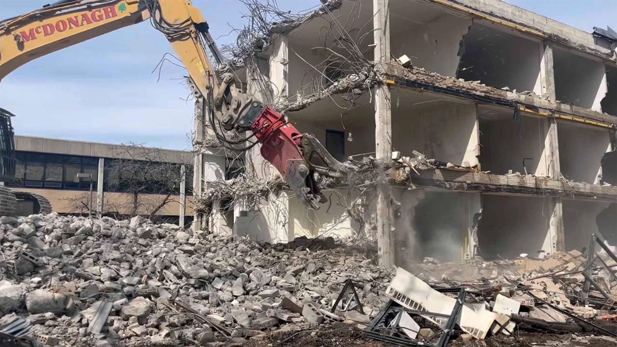 Excavator with hydraulic crusher attachment demolishing a multi-story concrete building, breaking reinforced walls and processing debris on a demolition jobsite.