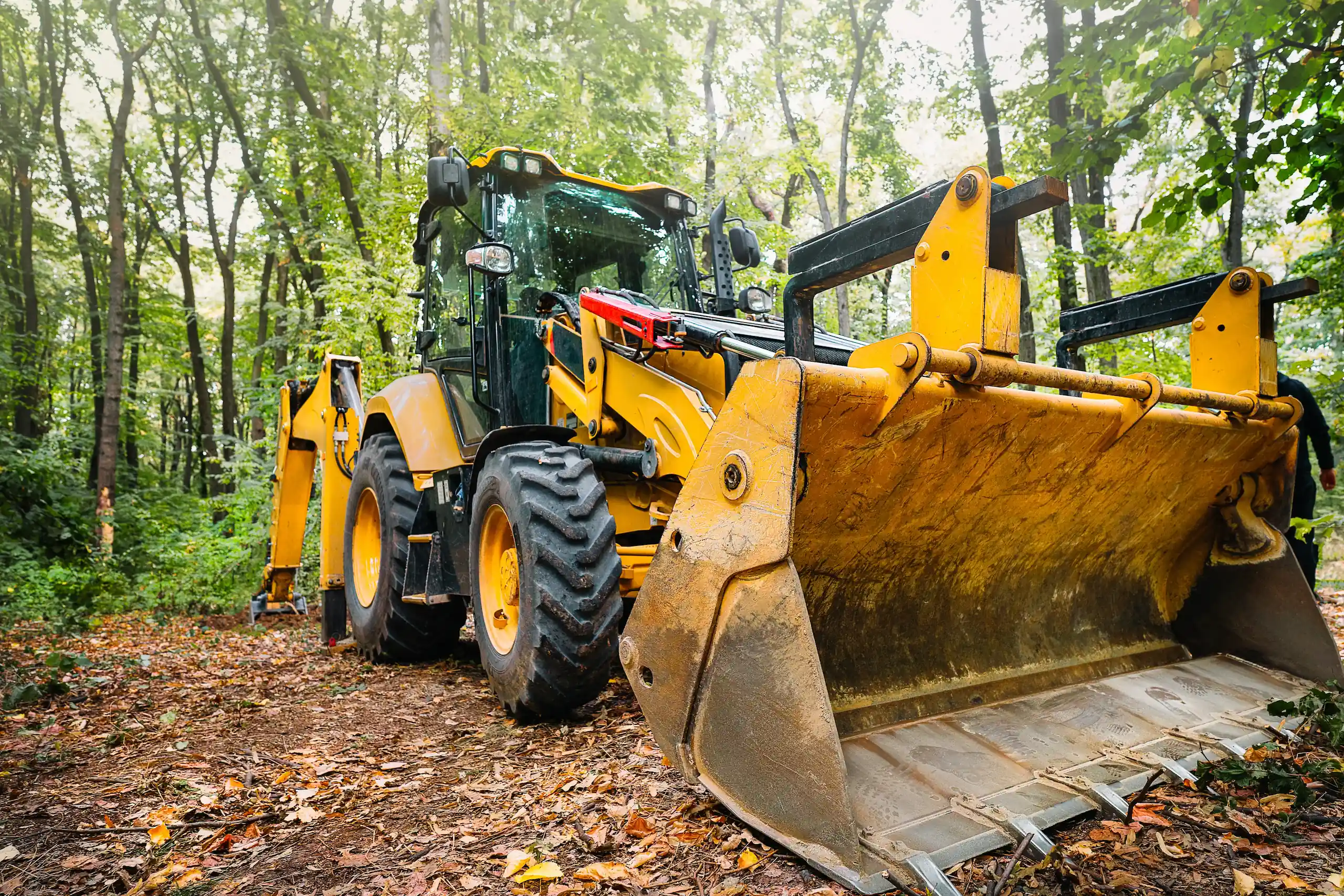 Large yellow backhoe loader clearing ground in a wooded jobsite, showing front bucket and heavy-duty tires used for land clearing and construction work.