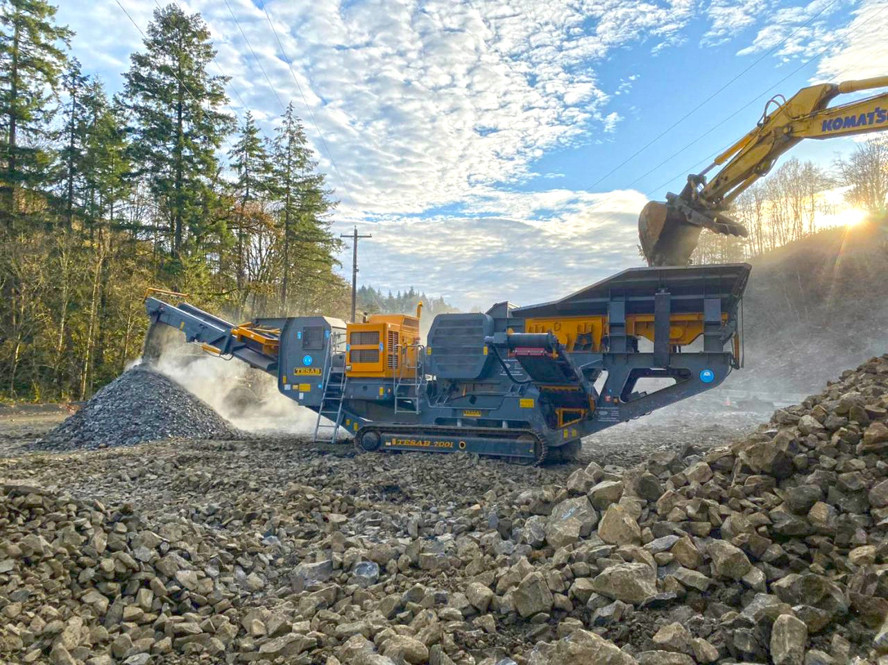 Tesab 700i mobile jaw crusher processing basalt as an excavator loads material, producing a large pile of crushed rock at a quarry site.