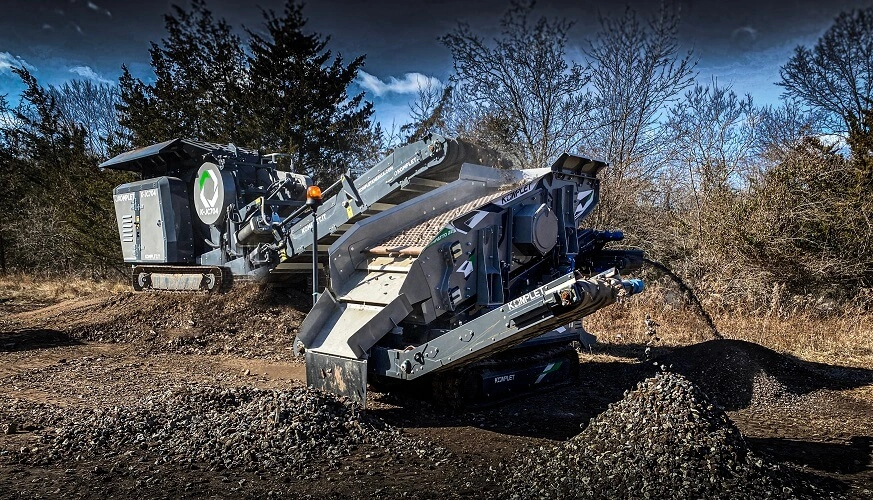 Komplet compact mobile crusher and screener working together on a jobsite, producing gravel piles during on-site recycling and material processing.
