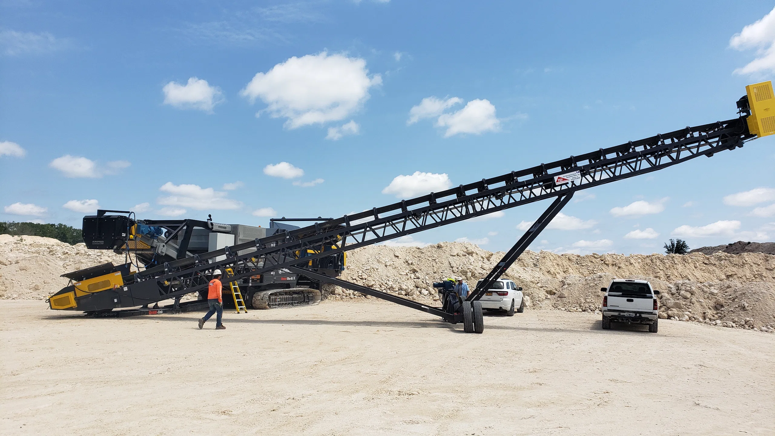 Tracked mobile conveyor system operating in a quarry with workers onsite, positioned to feed material into a crusher for aggregate production.