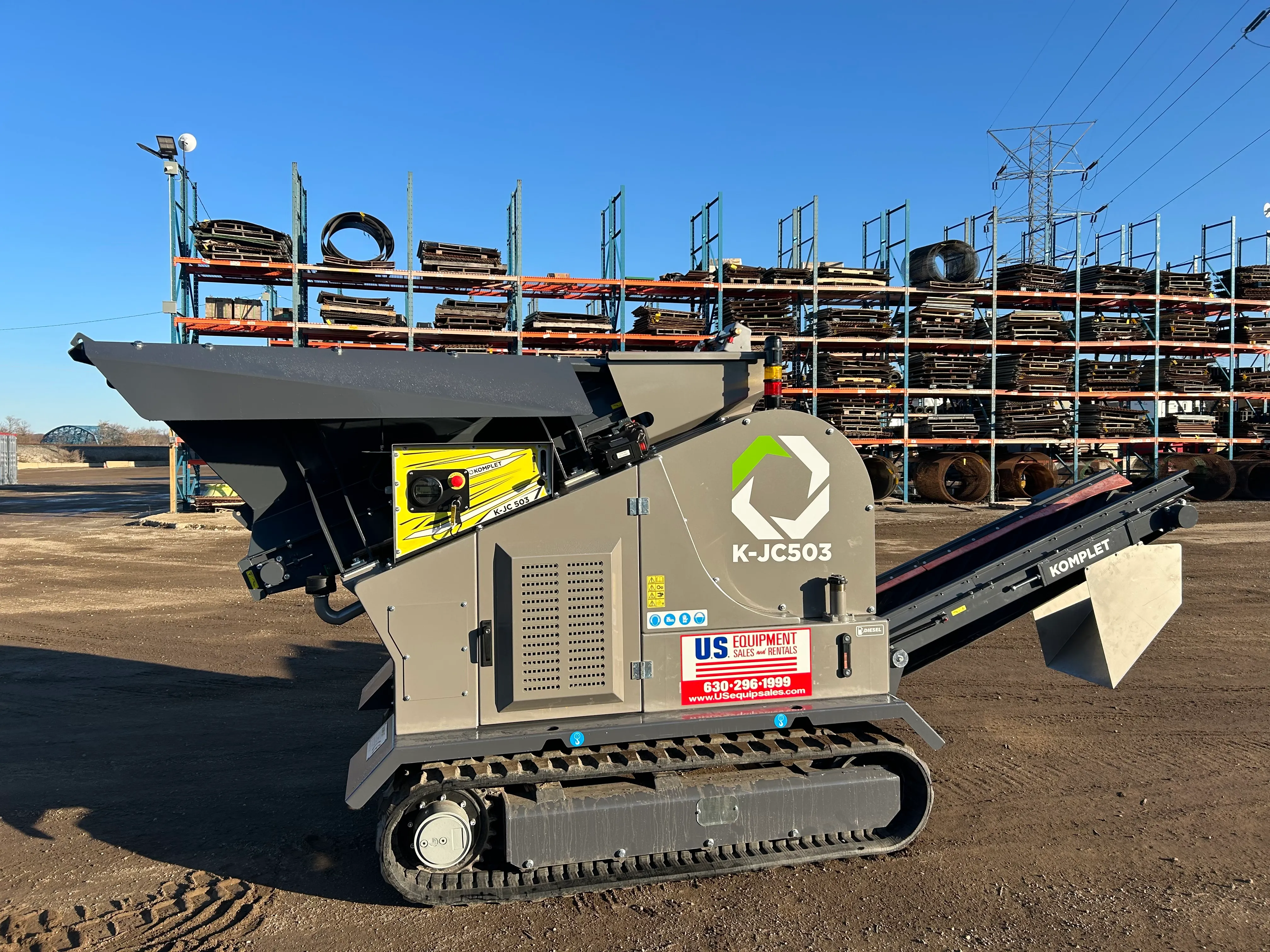 A compact, track-mounted Komplet K-JC503 jaw crusher with a gray body and yellow accents, featuring a large hopper and conveyor, branded with "US Equipment Sales."