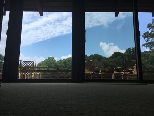 View through glass doors of an outdoor deck with wicker chairs and a small table under a partly cloudy blue sky with trees in the background.