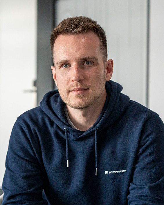 Portrait of a young man with short hair wearing a navy blue hoodie with the logo 'masyscon' seated indoors.
