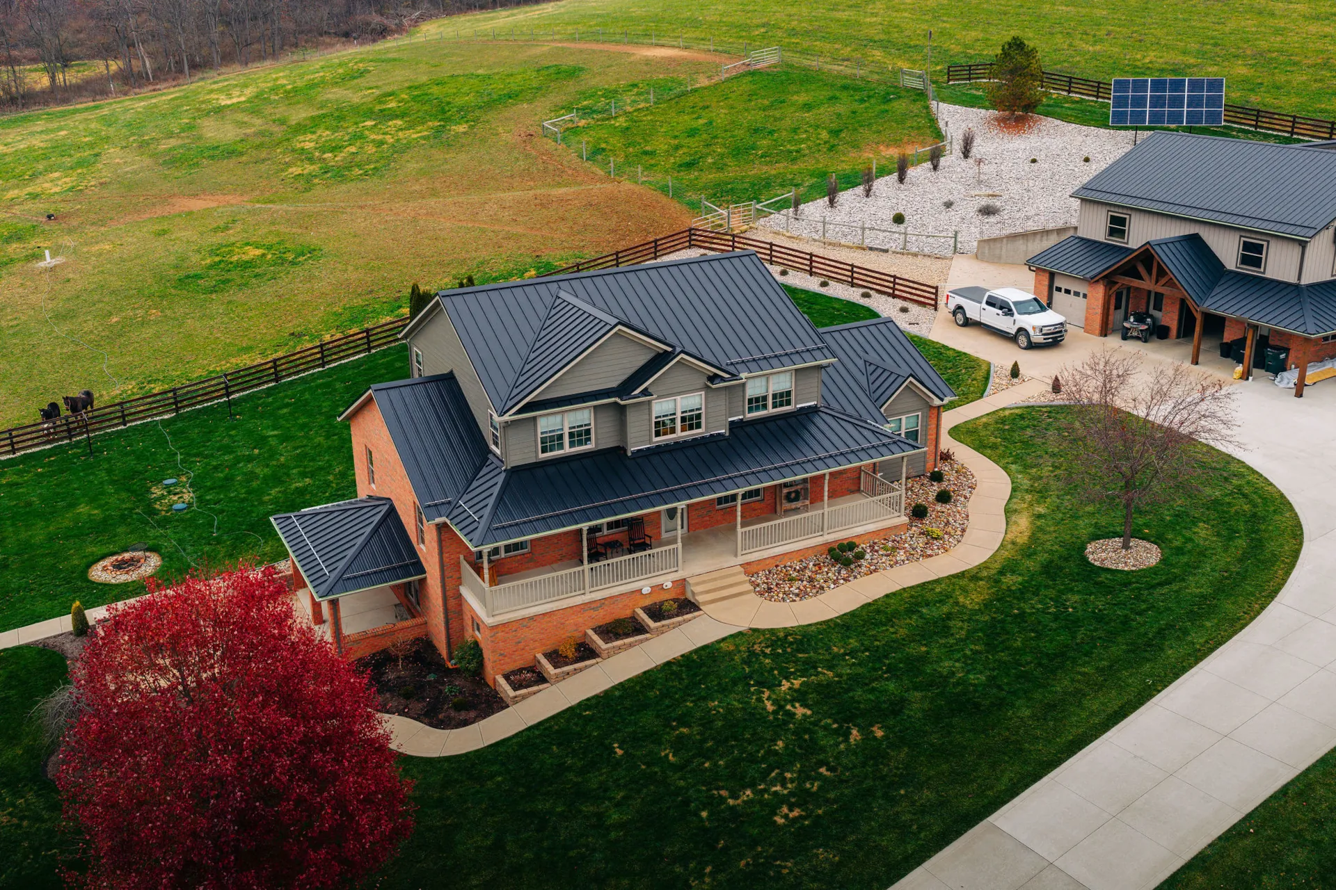 Aerial view of a brick home with a dark standing seam metal roof and wraparound porch, with a matching detached garage and solar panels in the background.