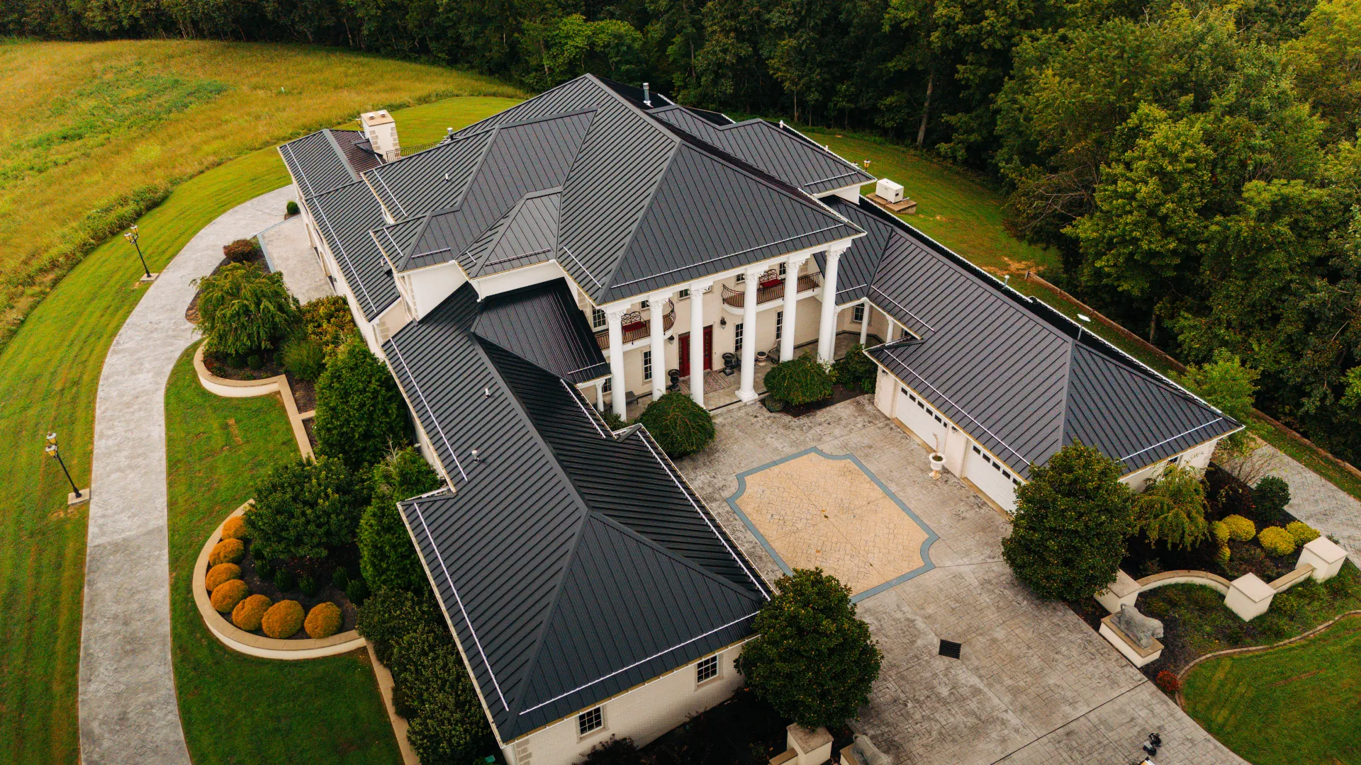 Aerial view of a large white home with a dark standing seam metal roof, multiple rooflines, and a columned front entrance surrounded by landscaped grounds.