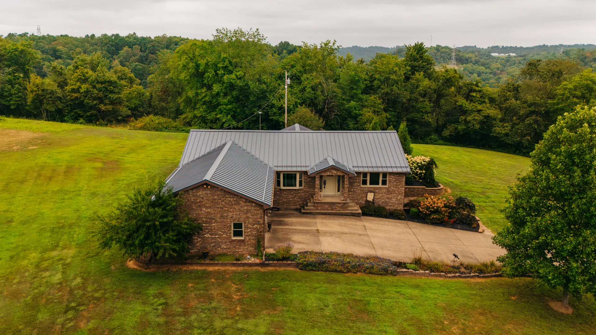 Aerial view of a brick home with a gray standing seam metal roof, front porch entry, and concrete driveway, surrounded by a large grassy yard and trees.