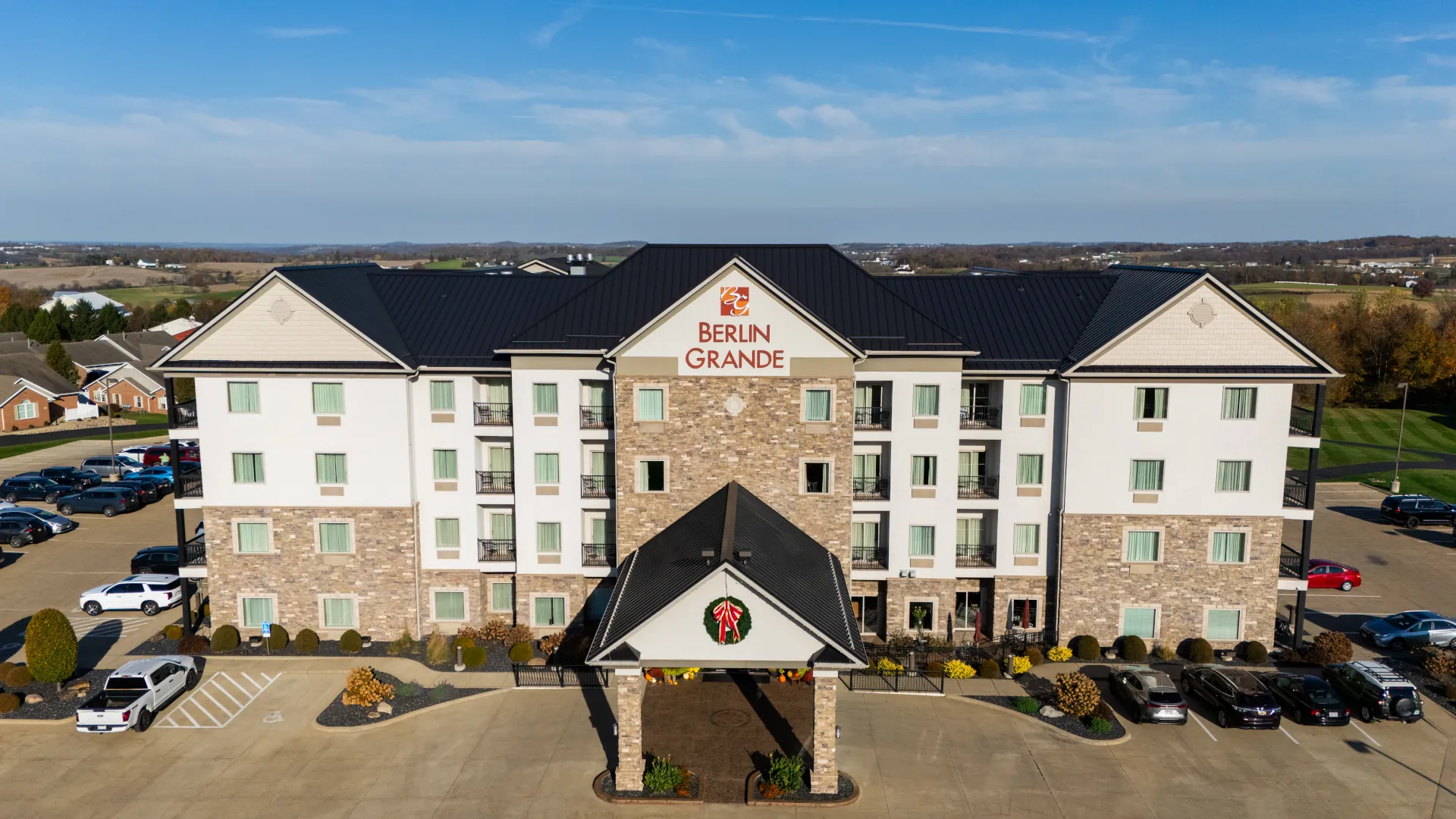 Aerial front view of the Berlin Grande hotel with a black standing seam metal roof, stone facade, and covered entrance, with parking lot in the foreground.