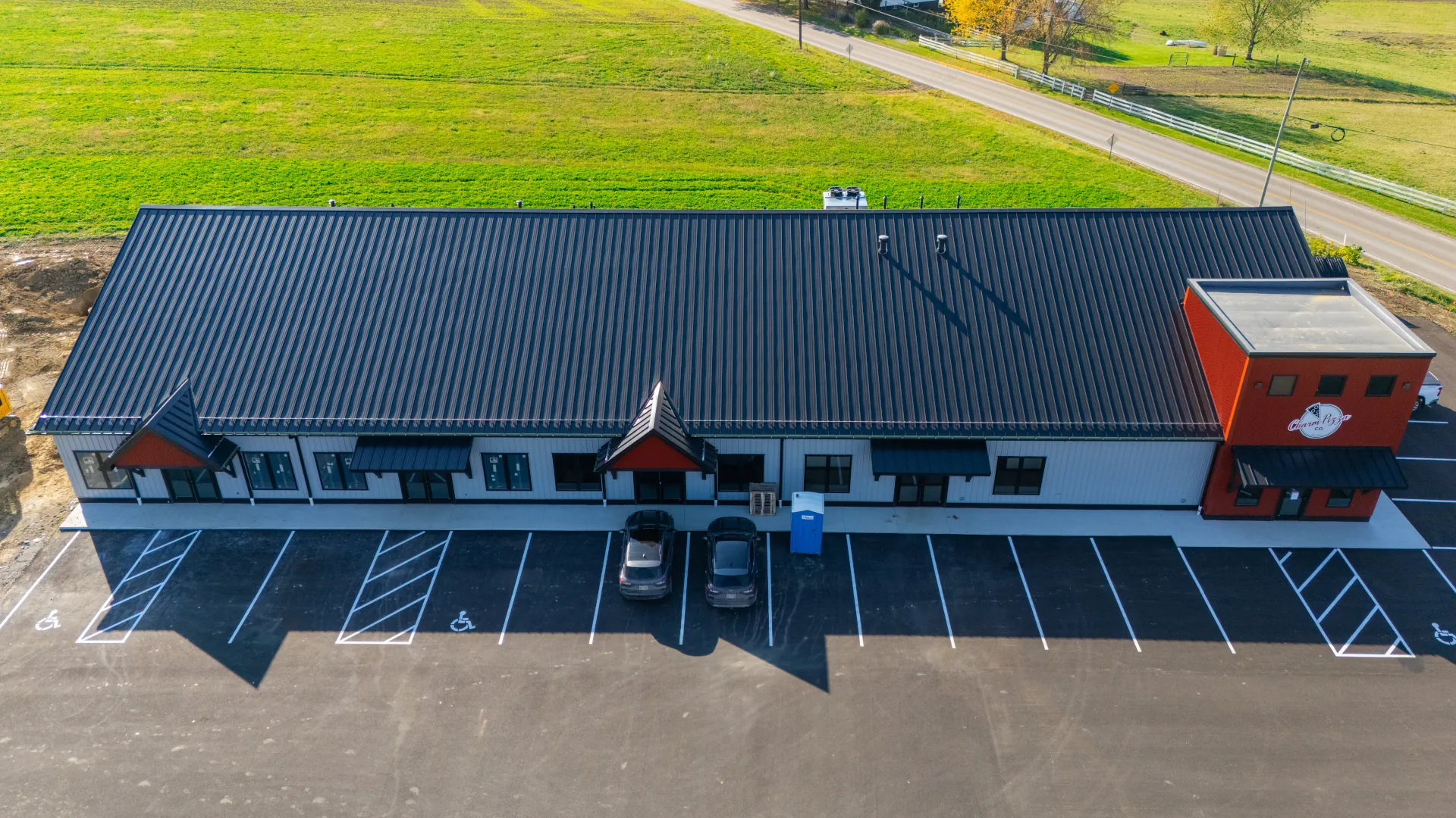 Aerial view of a long commercial building with a black standing seam metal roof, storefront entrances, and a parking lot in front, set beside a rural road and green fields.