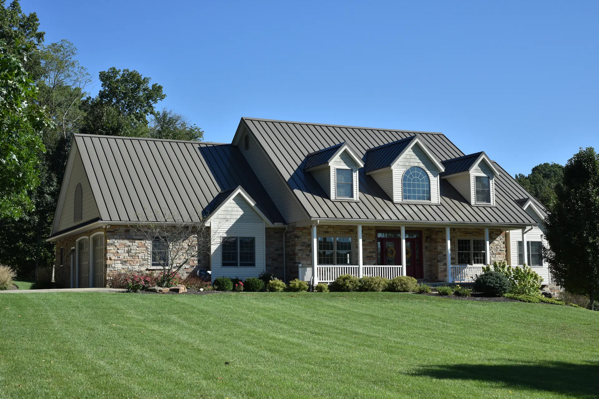 Large house with medium bronze standing seam metal roof, stone and white siding, dormer windows, front porch, and green lawn.