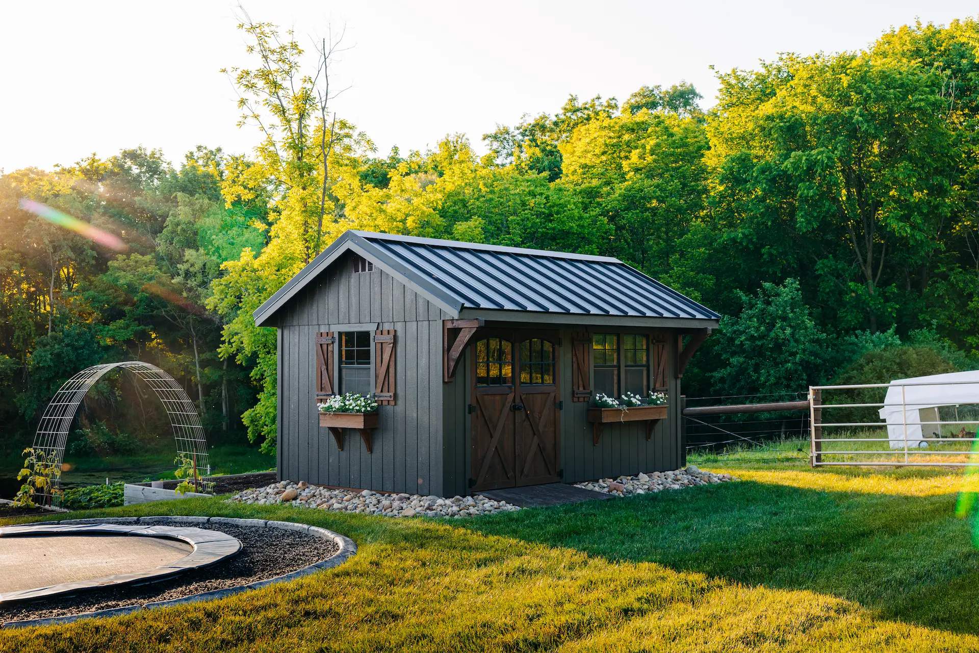 Gray garden shed with wooden doors, windows with flower boxes, and a dark bronze standing seam metal roof in green yard.