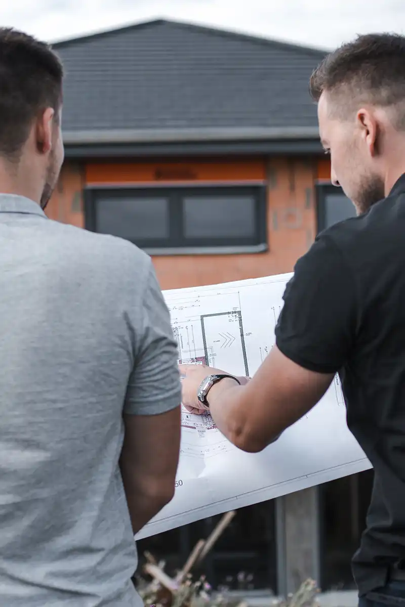 zwei Personen zeigen auf einen Bauplan mit einem Haus im Hintergrund