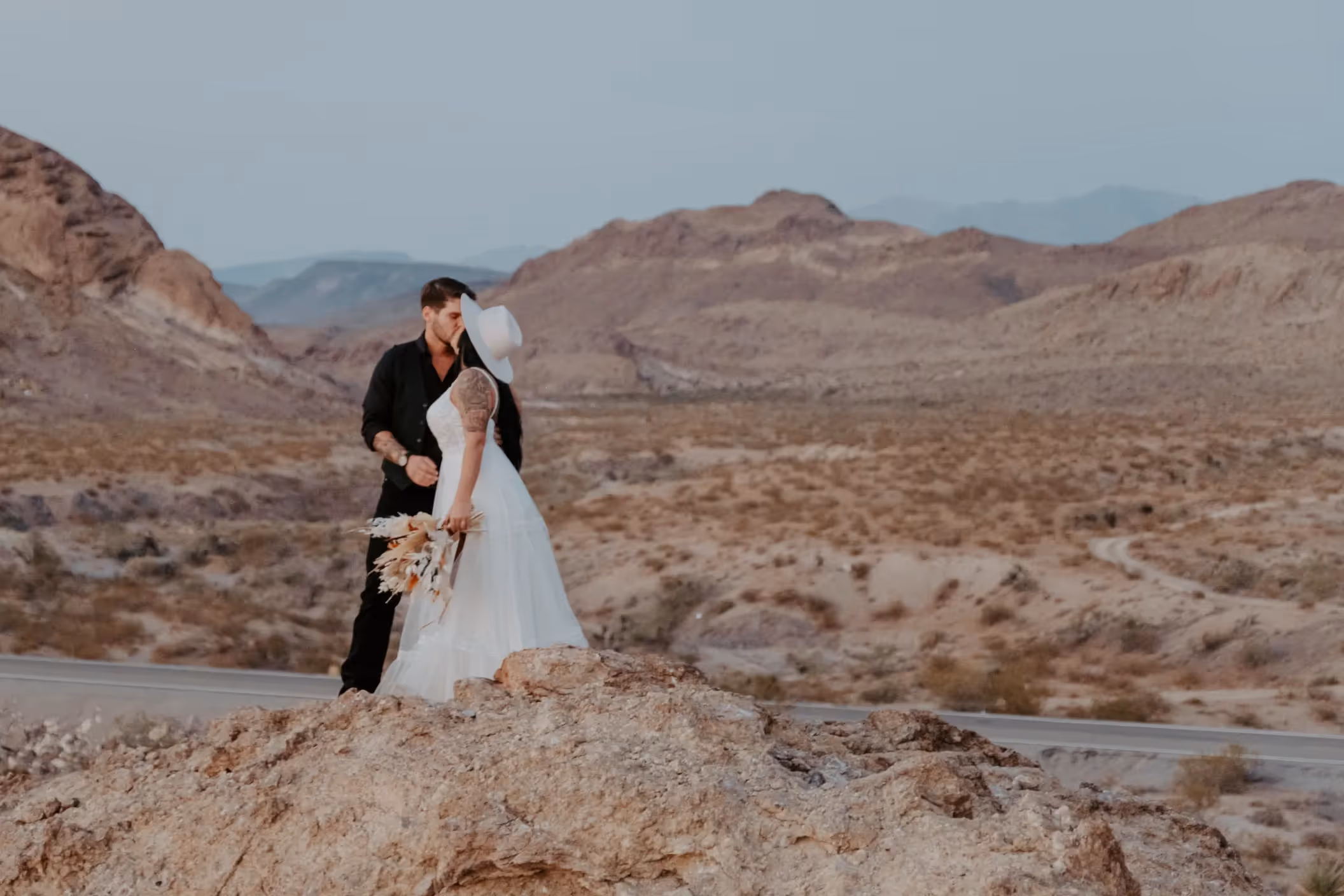 Hannah and Patrick, bride in white dress with cowboy hat kissing groom on rocky overlook with expansive mountain views