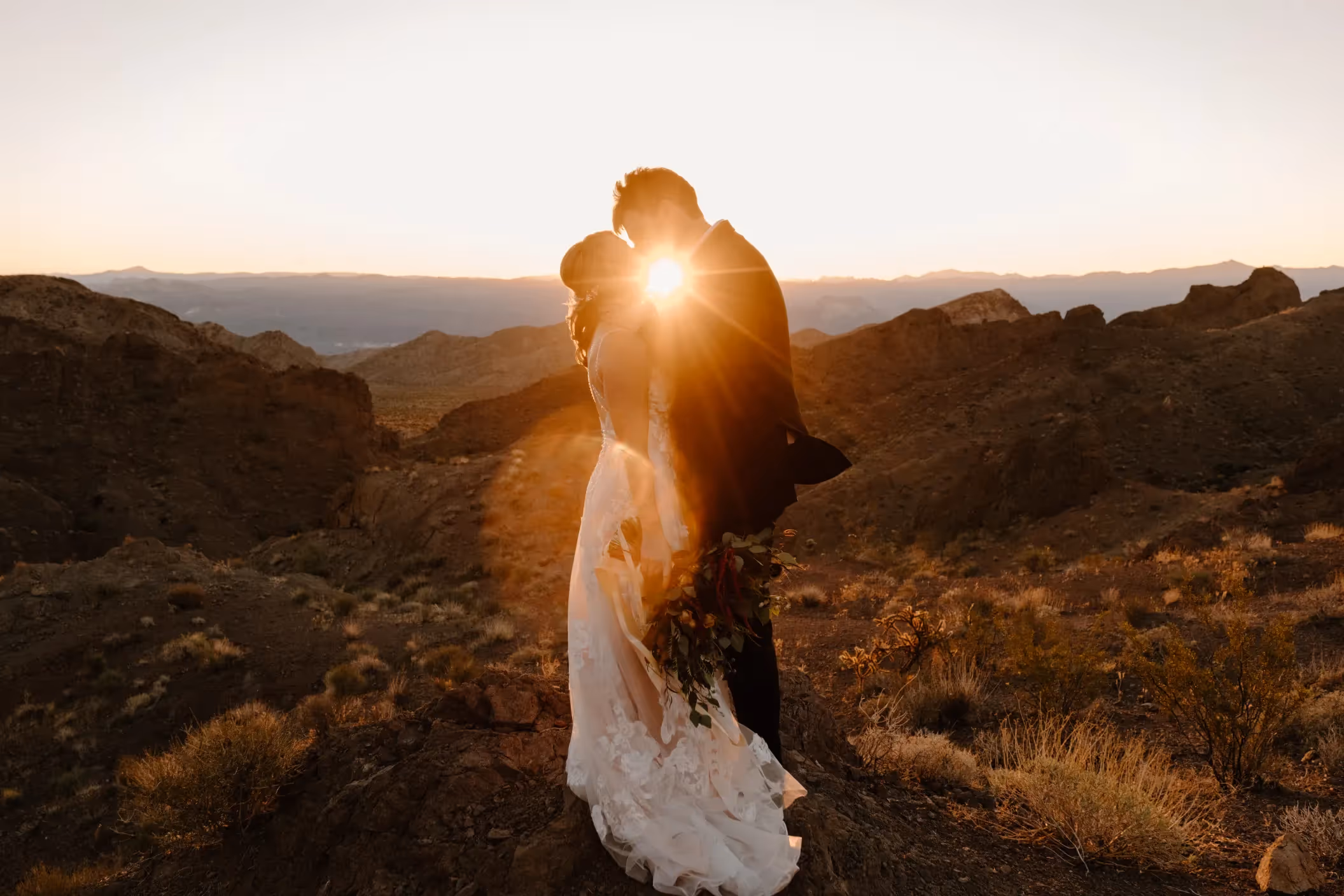 Breathtaking sunset elopement of Amy and Bill in Las Vegas desert, silhouette of couple with sun peeking through, surrounded by rugged mountain landscape