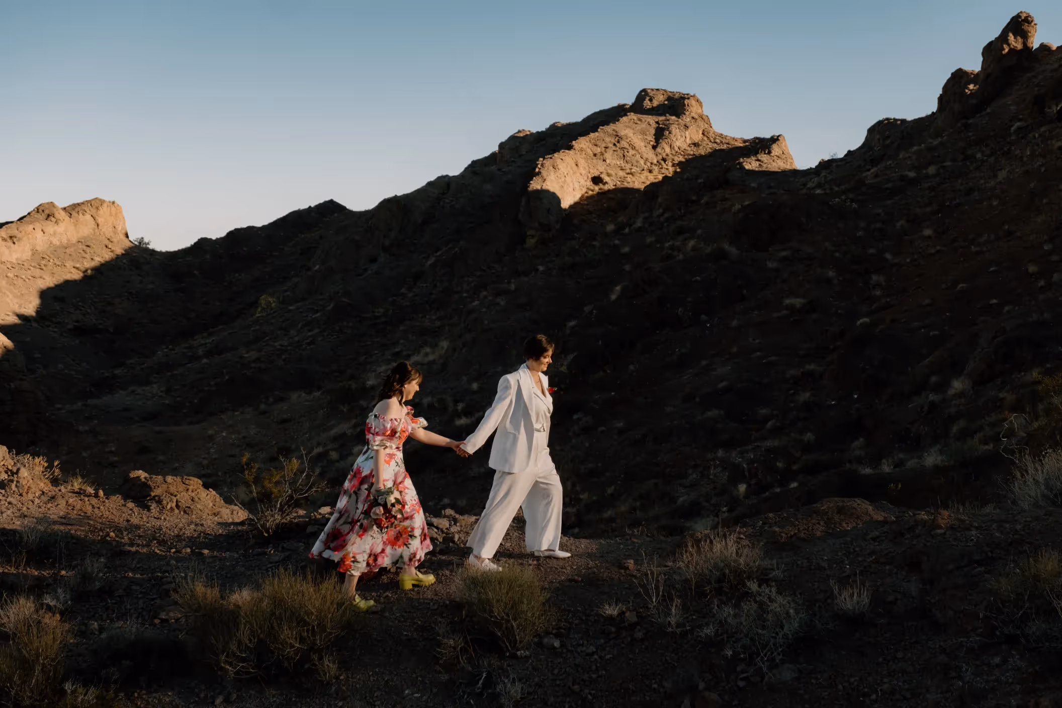 Desert wedding of Rachel and Emma, brides in contrasting outfits walking hand-in-hand against dramatic rocky landscape at sunset