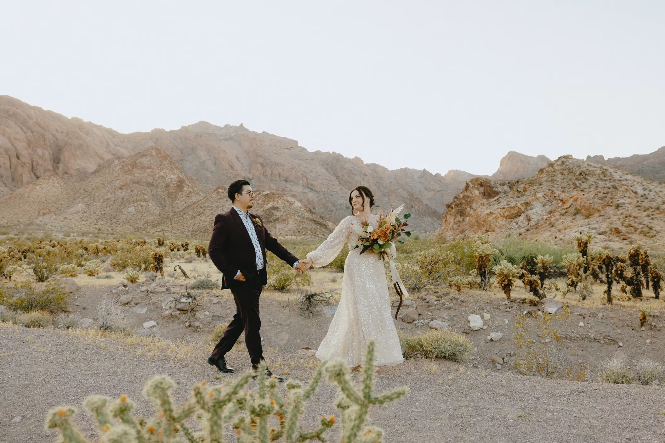 Carla and Brian's desert elopement at Nelson Ghost Town, couple strolling hand-in-hand among cacti with rugged mountains in background