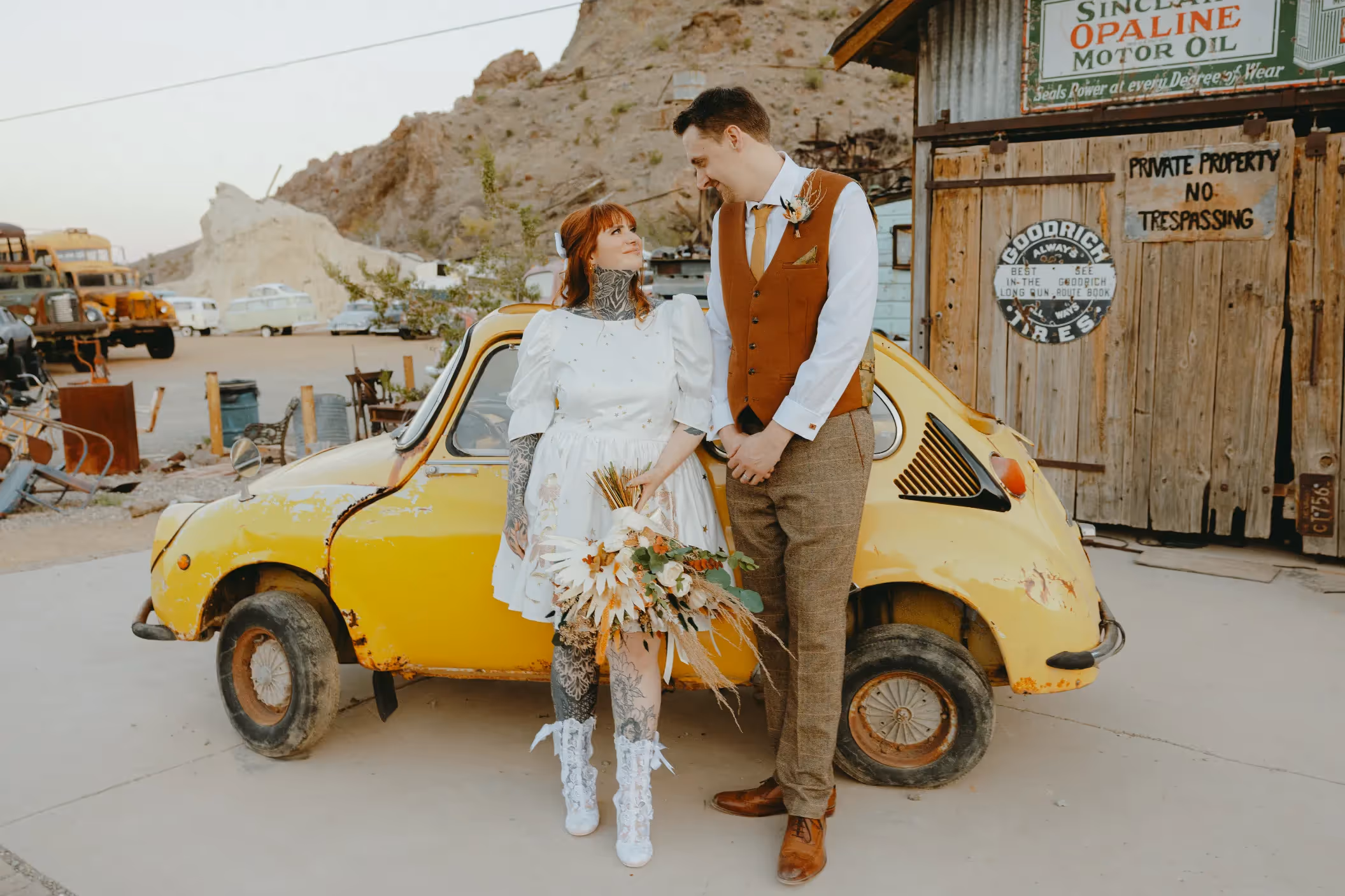 Area wedding photo of Maddie and Sam at Nelson Ghost Town, tattooed bride in short dress and groom by vintage yellow car with rustic buildings