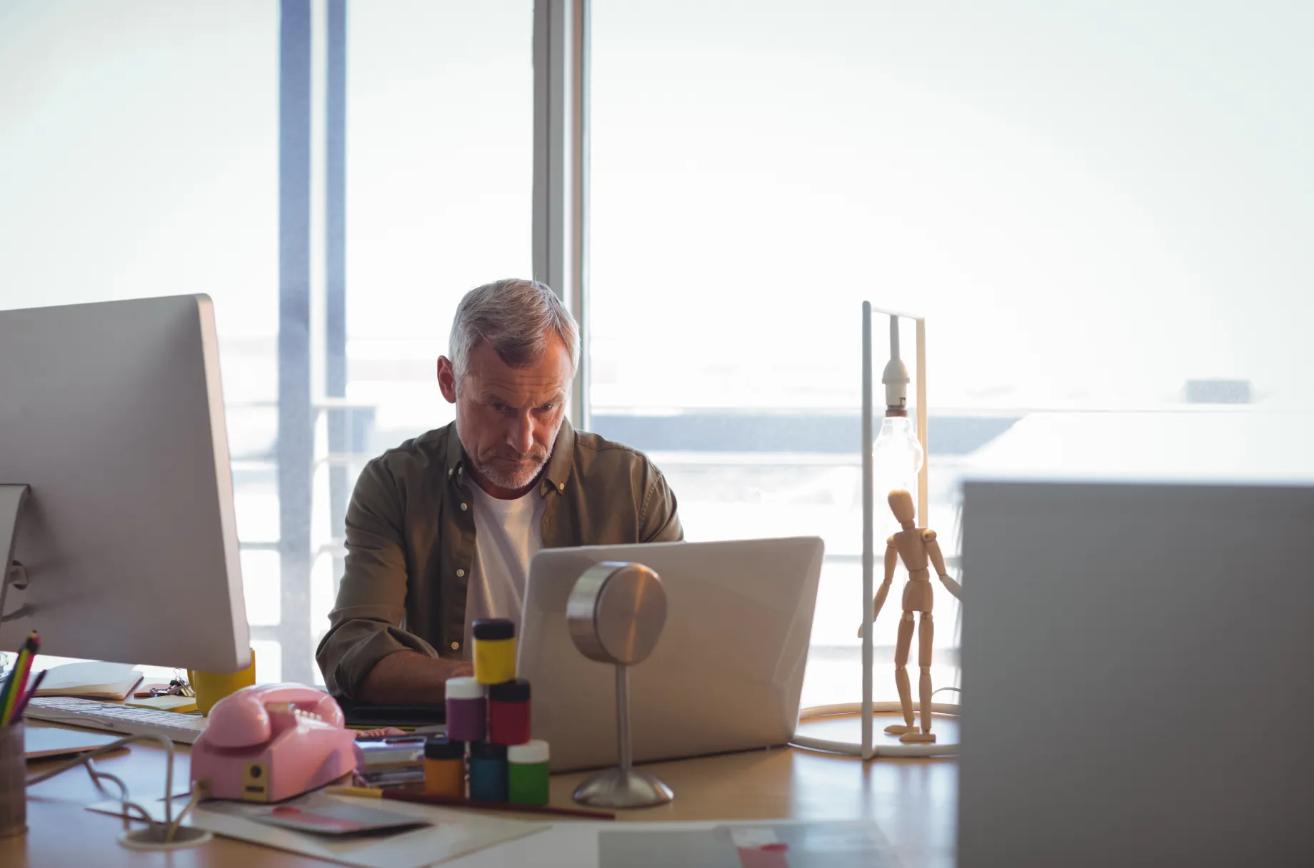 Man with gray hair working on a laptop at a desk with art supplies and a wooden mannequin figure.