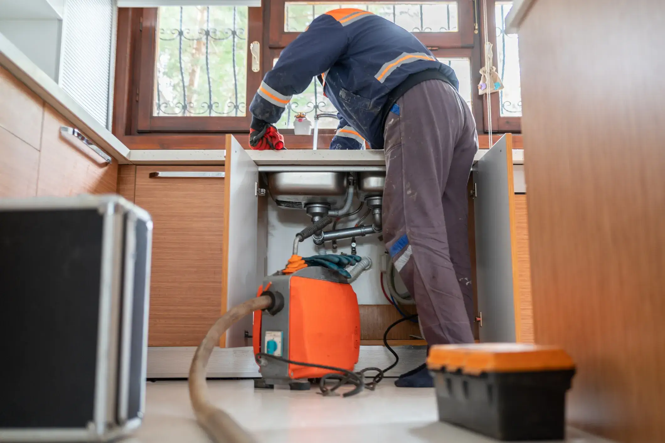 Plumber in blue jacket using vacuum pump under kitchen sink with toolbox and tools nearby