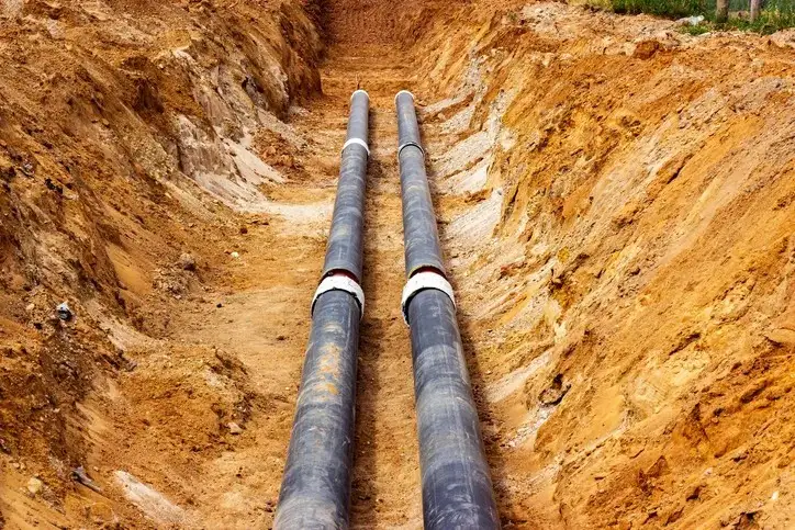 Two large, black pipes laid side-by-side in a deep trench of brown dirt. The trench is surrounded by more loose dirt.