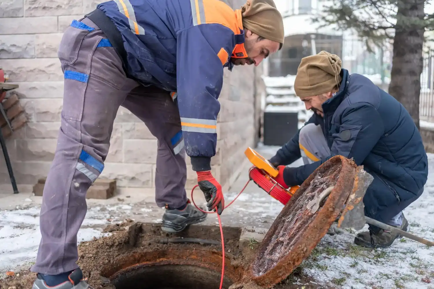 Two utility workers, dressed in warm winter gear, are inspecting an open manhole on a snowy or frosty day. 
