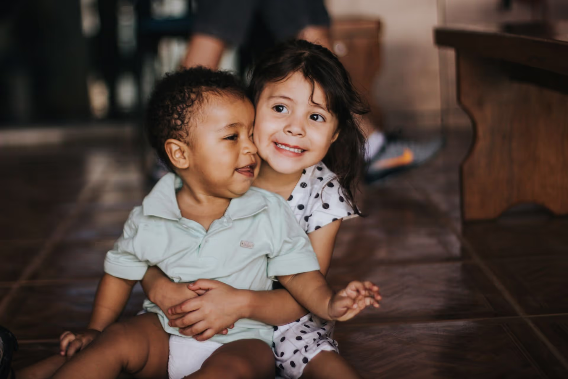 A young child in a light green polo sits on the floor, being embraced by another child in a polka dot dress, against a homey backdrop.