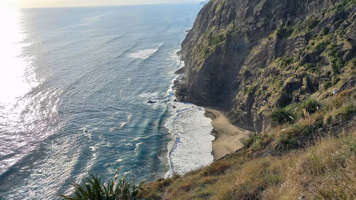 Cliffside view of a sunlit ocean with waves crashing onto a secluded sandy beach. The rocky cliff is lush with greenery, exuding a serene atmosphere.