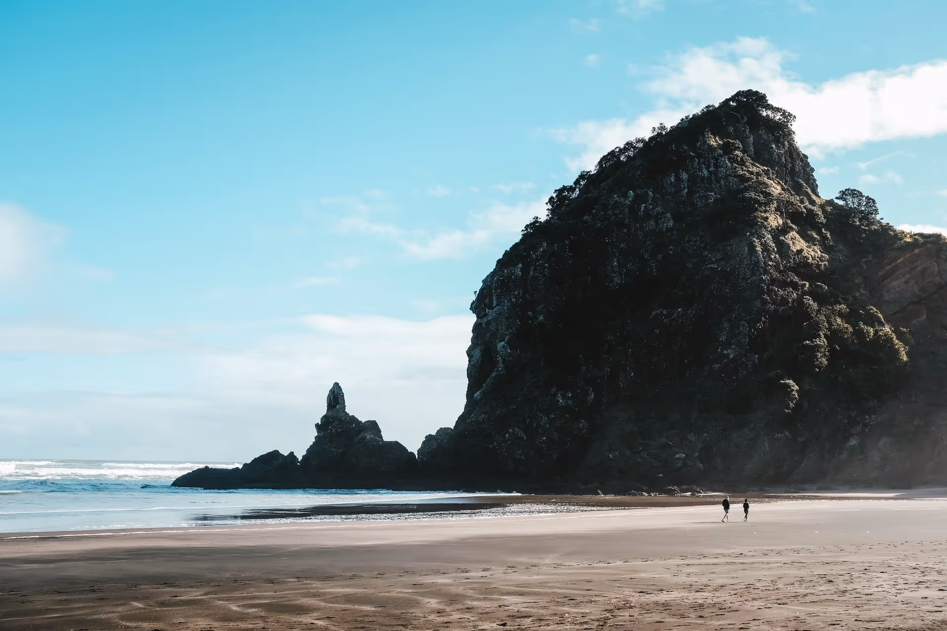 Wide beach with towering rock formations under a bright blue sky. Two people walk along the sand, conveying a sense of calm and serenity.