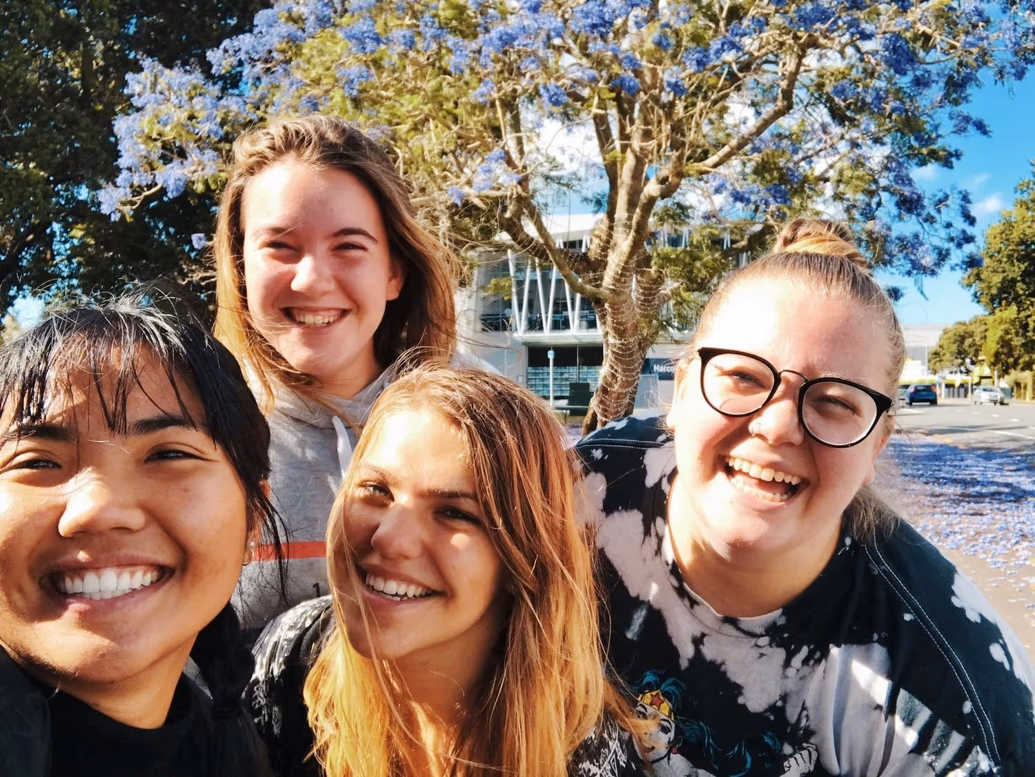 Four friends smiling widely under a blooming jacaranda tree with purple flowers. The bright day and their joyful expressions convey happiness.