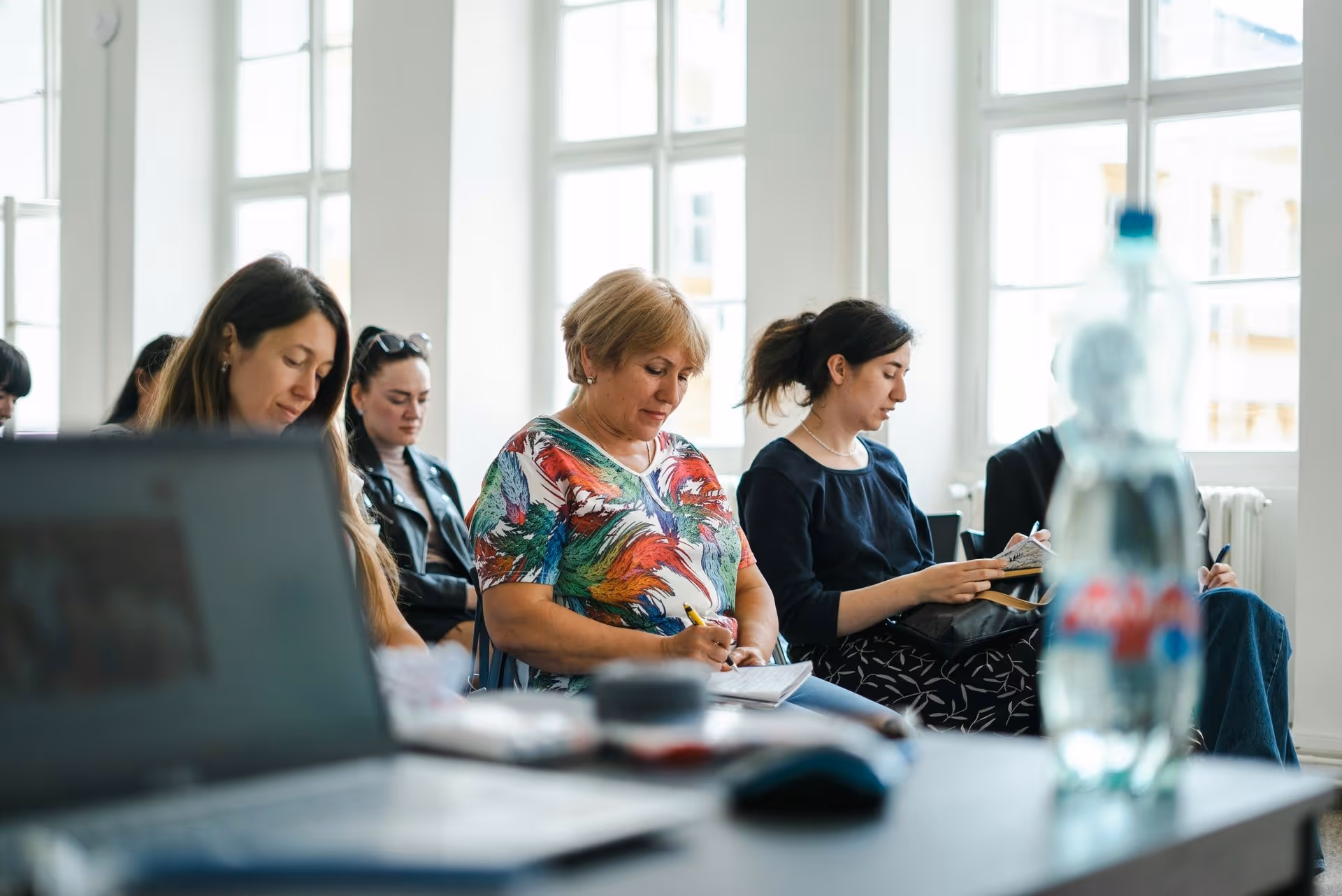 A group of women sit in a bright room with large windows, writing in notebooks. A blurred water bottle and laptop are visible in the foreground.