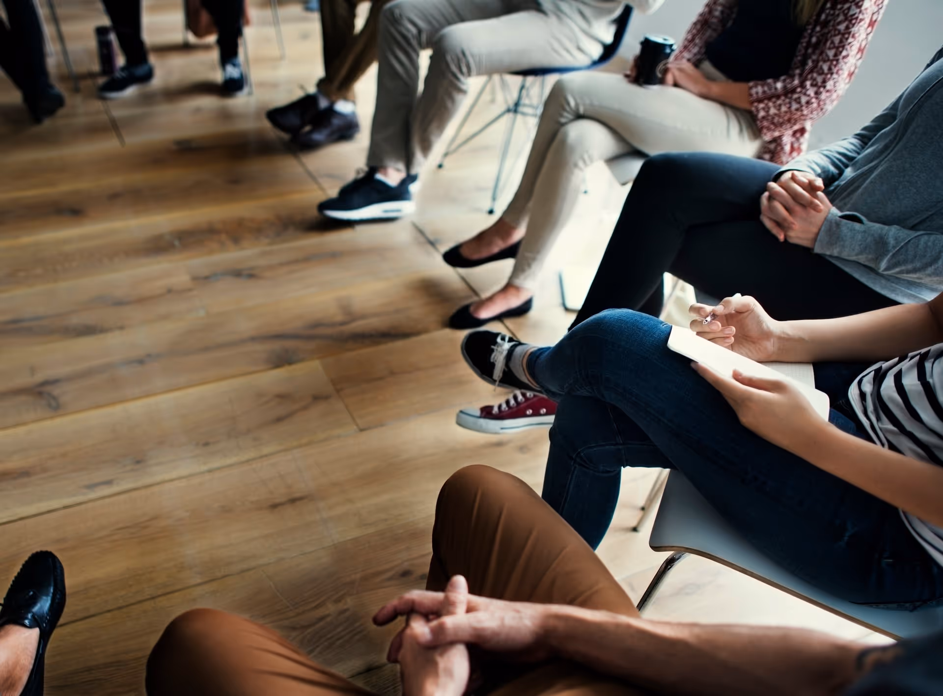 A diverse group of people seated in a meeting, some writing notes, with a wooden floor in the background, creating a collaborative atmosphere.