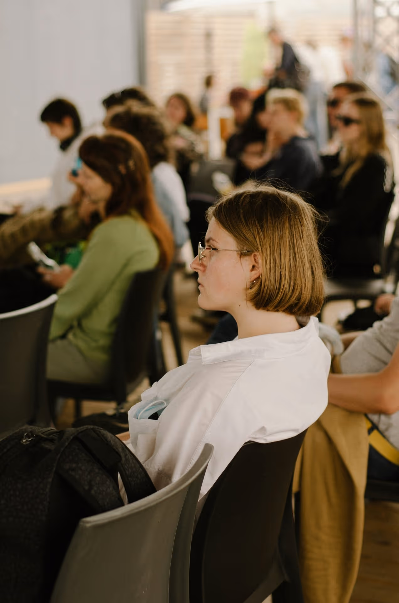 A group of people seated in a row, engaged in conversation or focused on their phones in a casual indoor setting.