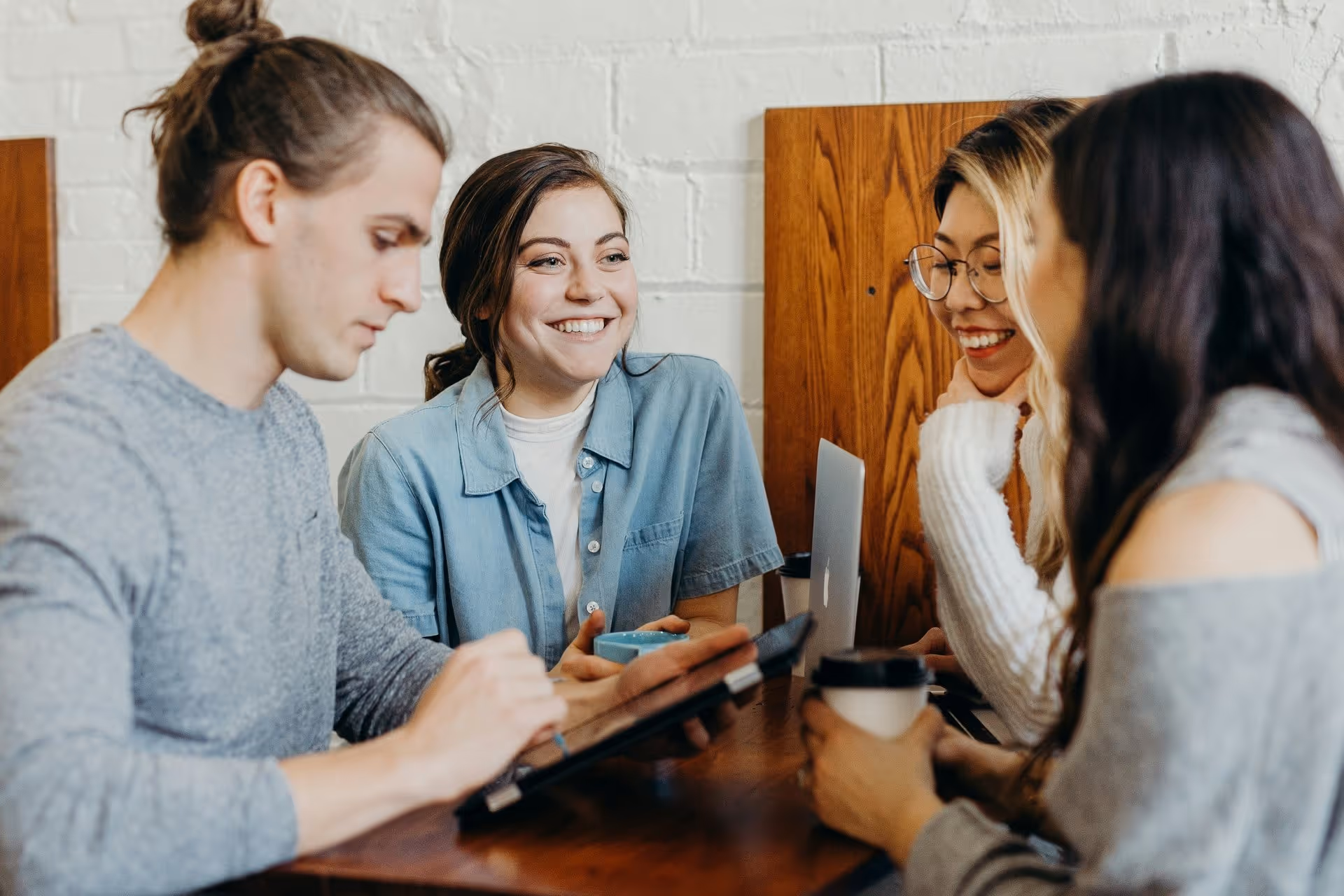 A group of four people engaged in discussion at a table, with a laptop and coffee cups among them in a cozy indoor setting.