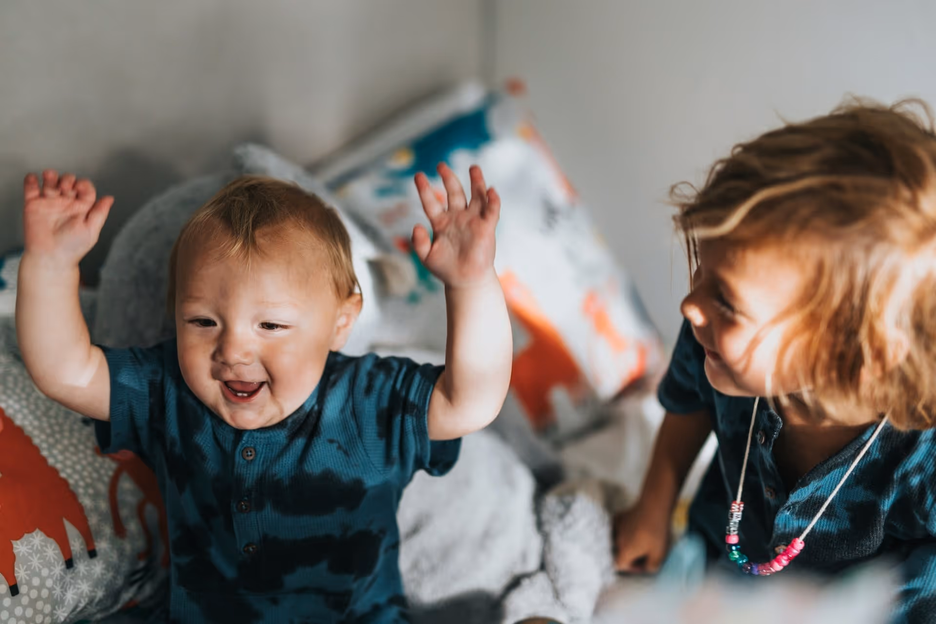 Two young children play together indoors, one raising their arms in joy, surrounded by colorful pillows and a cozy blanket.