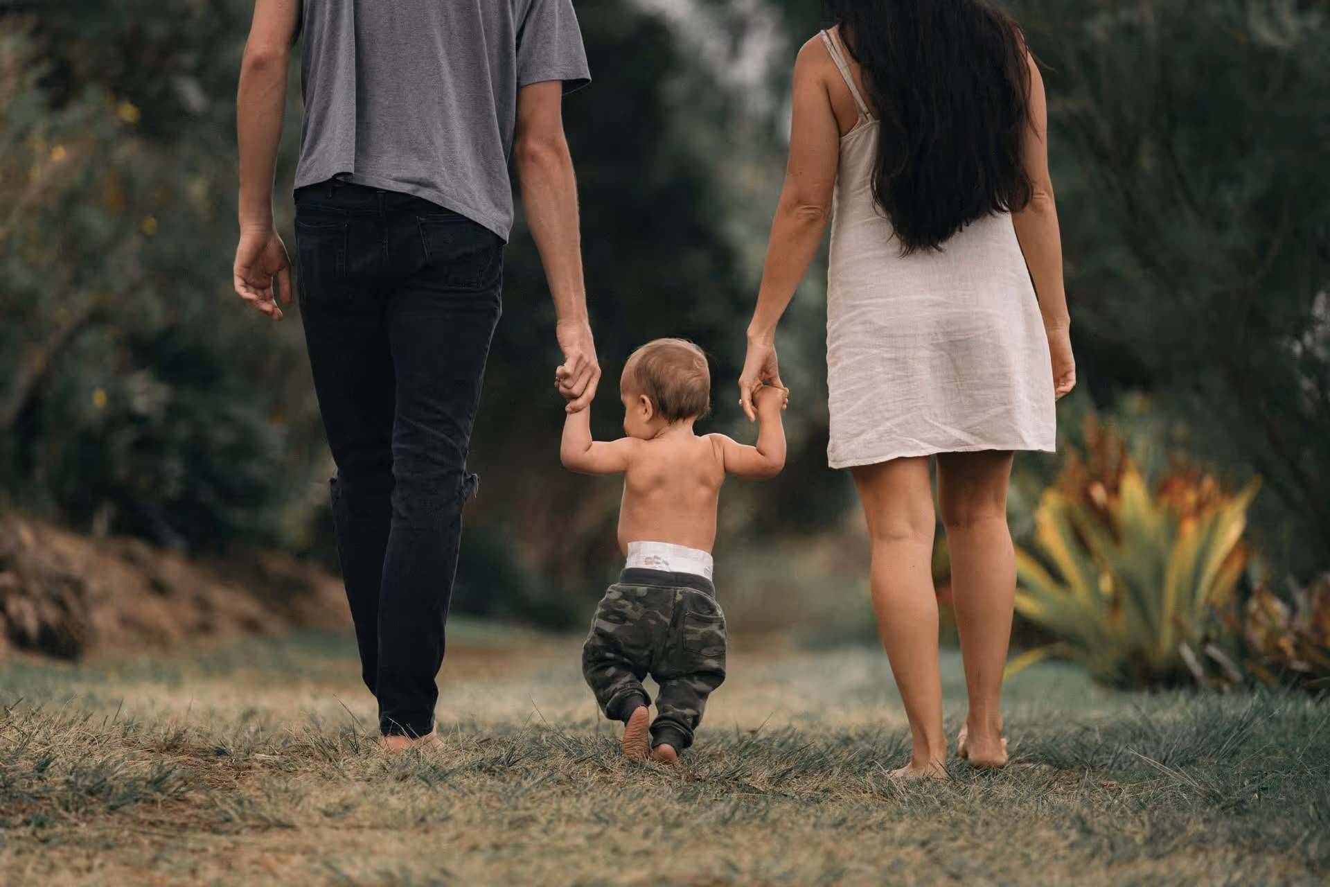 A couple holds hands with their toddler, walking barefoot on a grassy path surrounded by greenery, enjoying a family moment together.