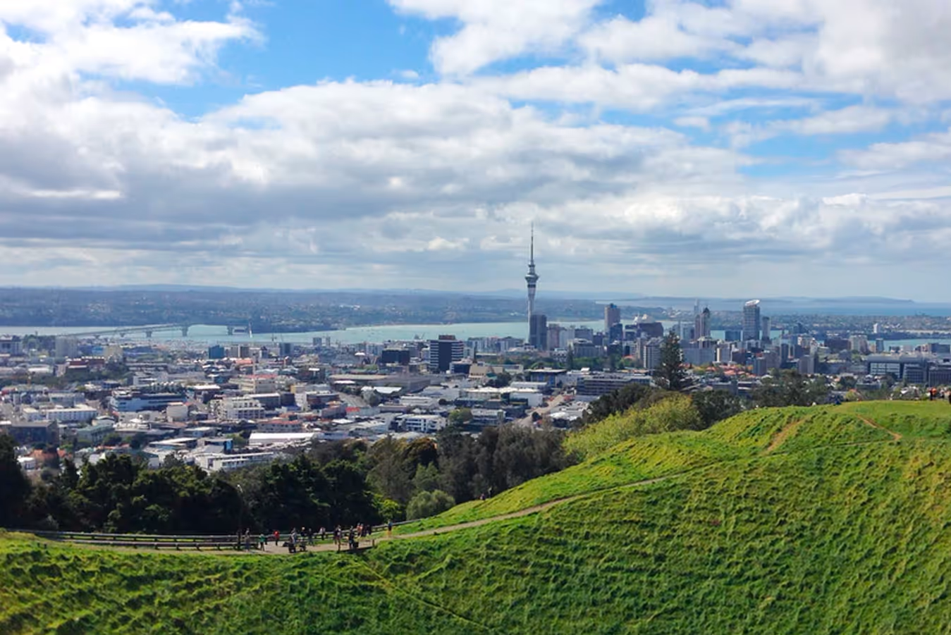 A panoramic view of Auckland, New Zealand, featuring the Sky Tower, lush green hills, and a vibrant urban skyline under partly cloudy skies.
