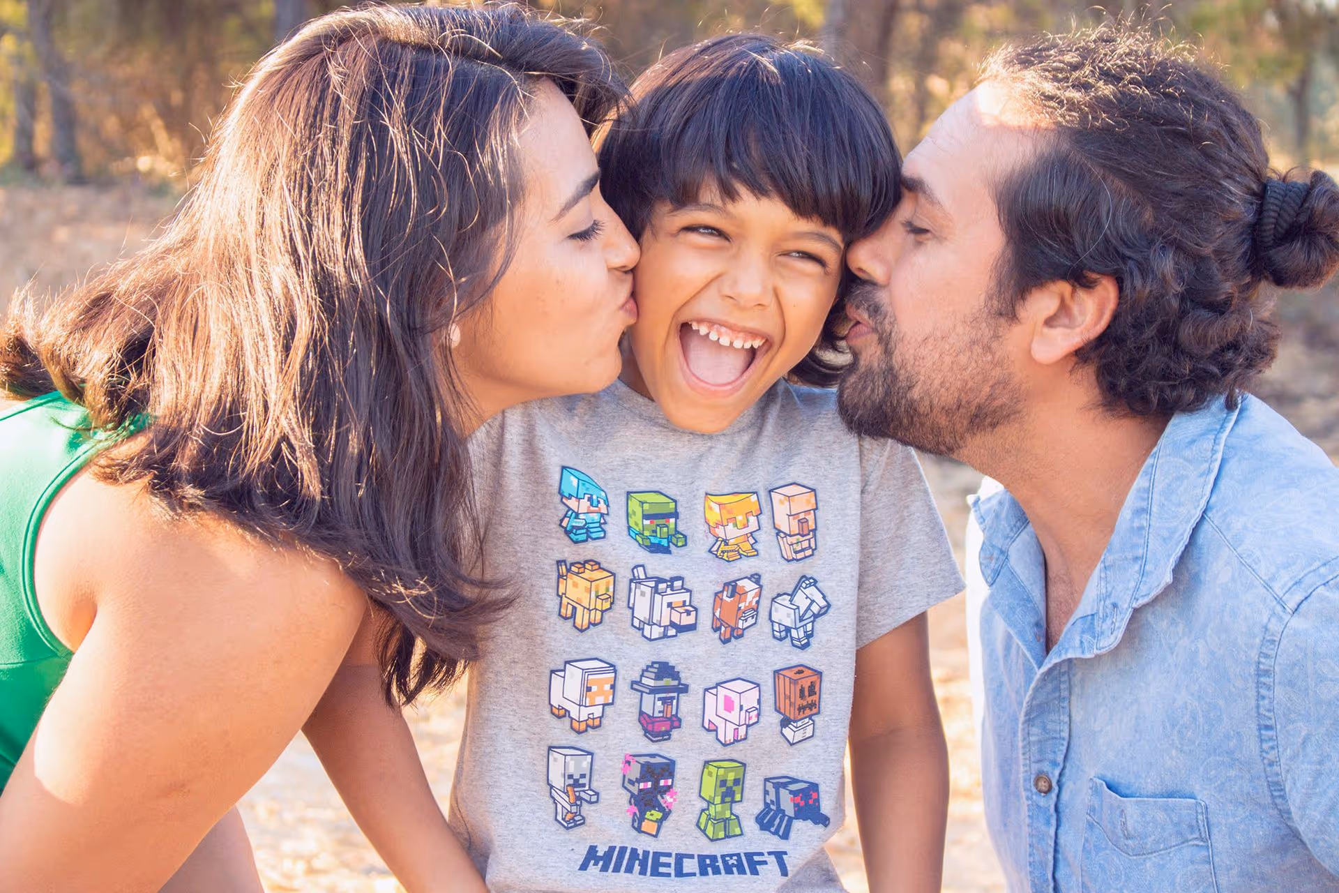 A woman and a man lean in towards a child wearing a Minecraft-themed shirt, surrounded by a natural outdoor setting.